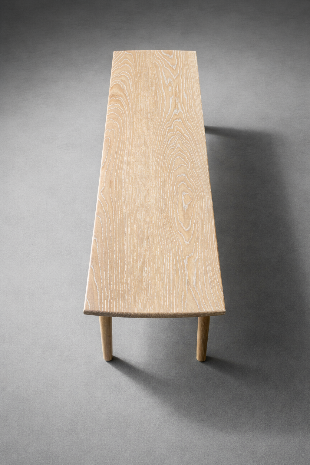 A wooden bench with a light wood surface and rounded legs, casting a shadow on a gray background.