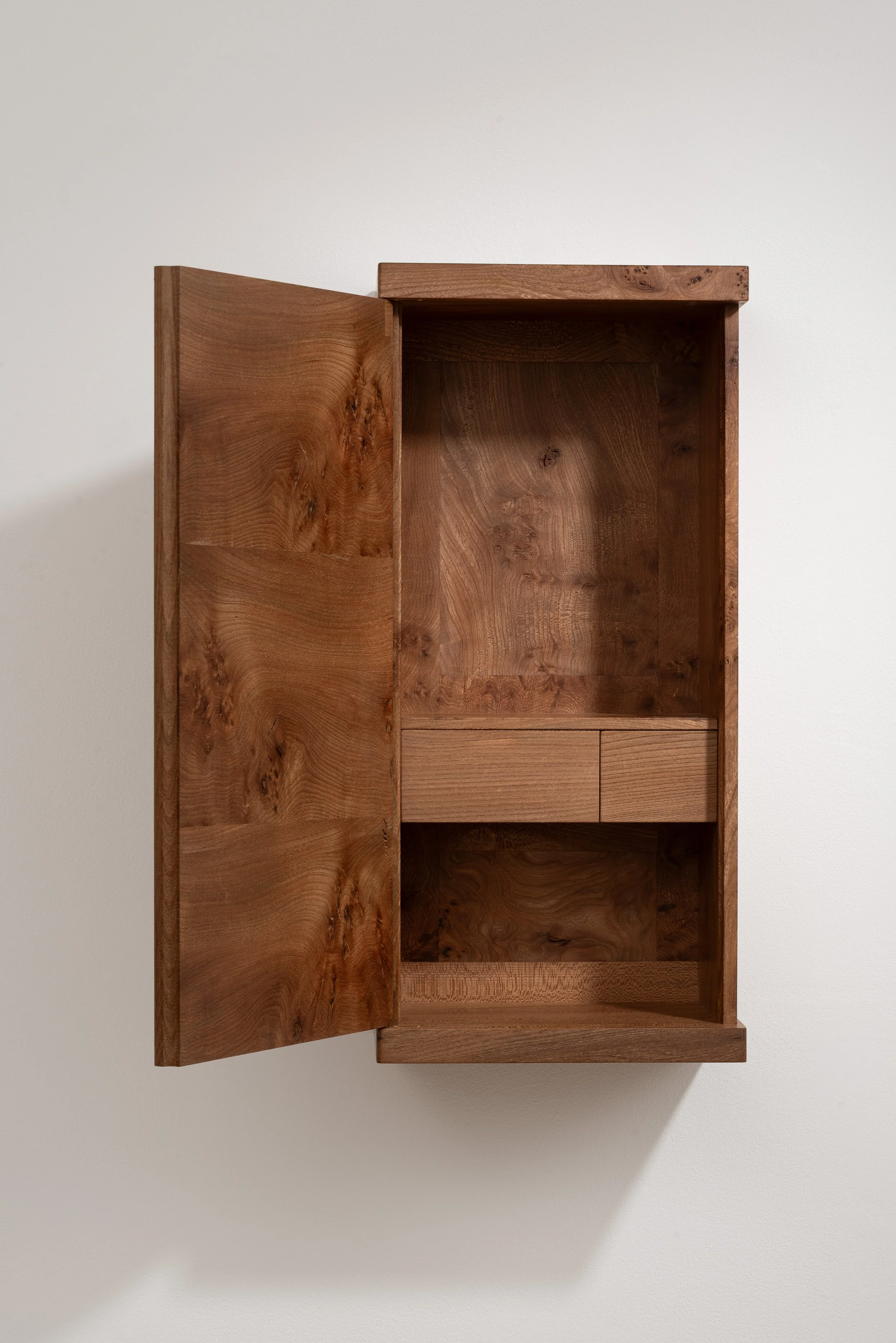 Empty wooden cabinet mounted on a white wall, with an open door revealing two small drawers inside.