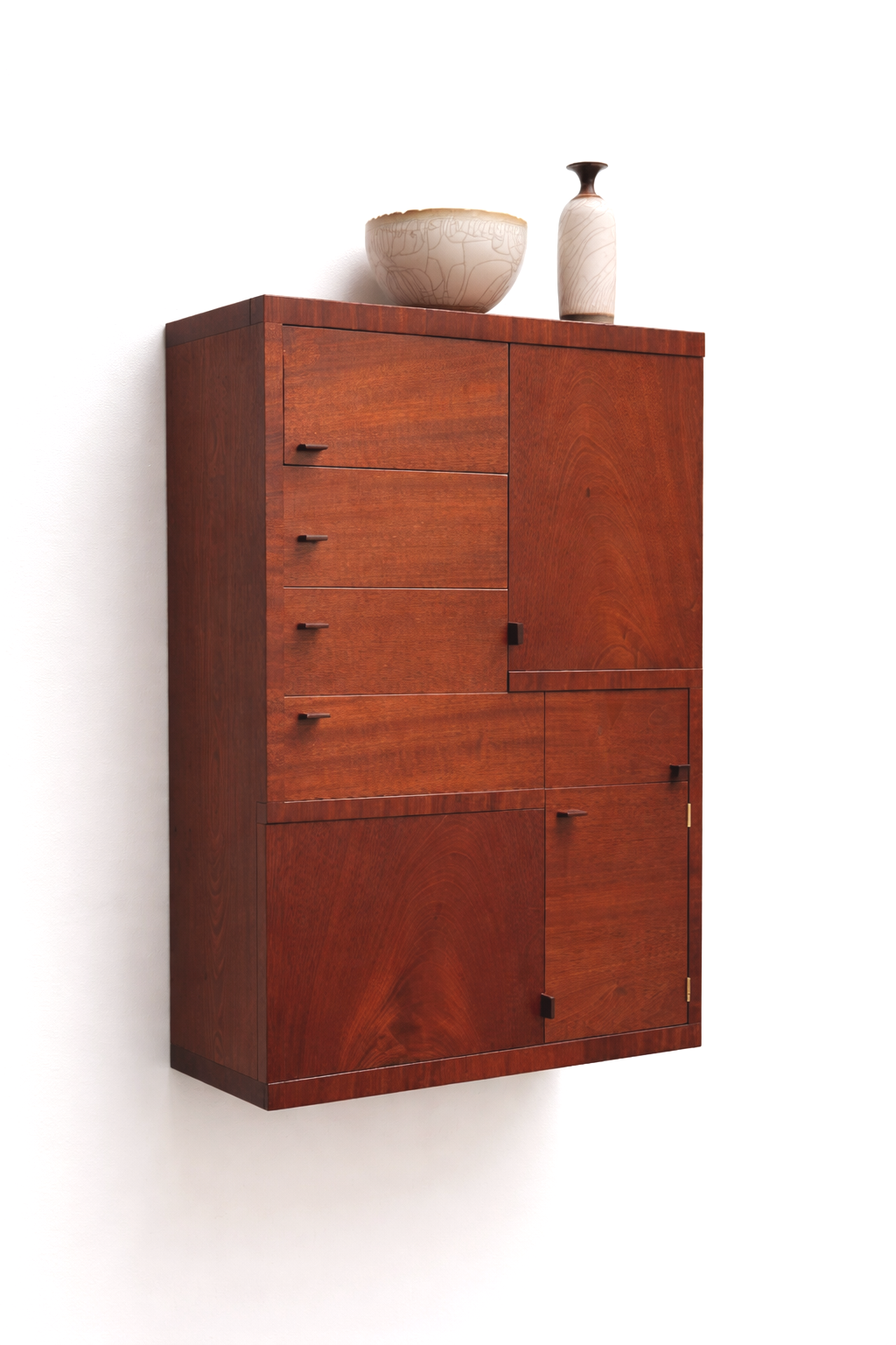 A tall wooden cabinet with drawers and doors, mounted on a white wall, topped with two ceramic vases and a bowl.