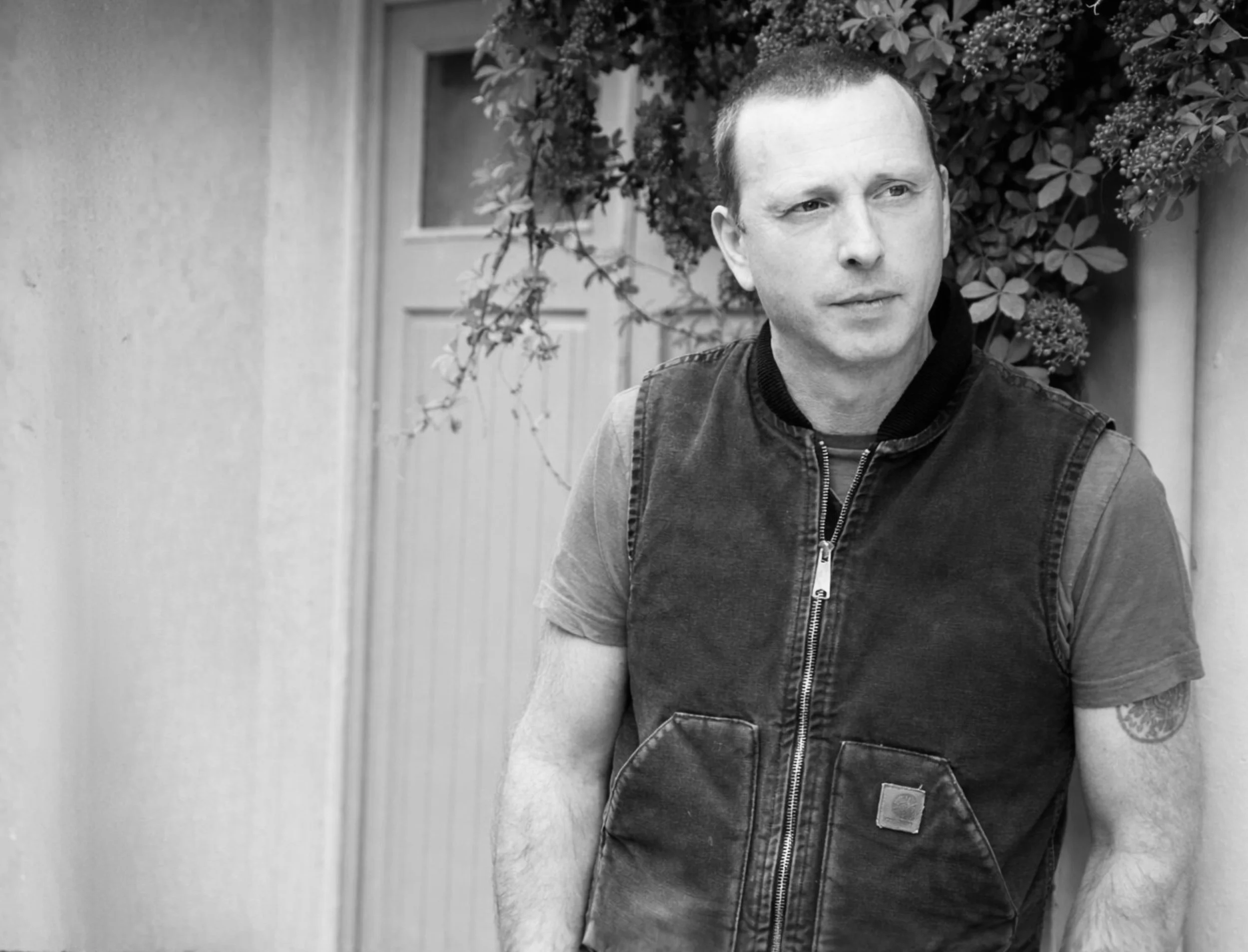 A man stands outdoors near a door and plants, wearing a sleeveless jacket over a t-shirt, looking to the side with a contemplative expression.