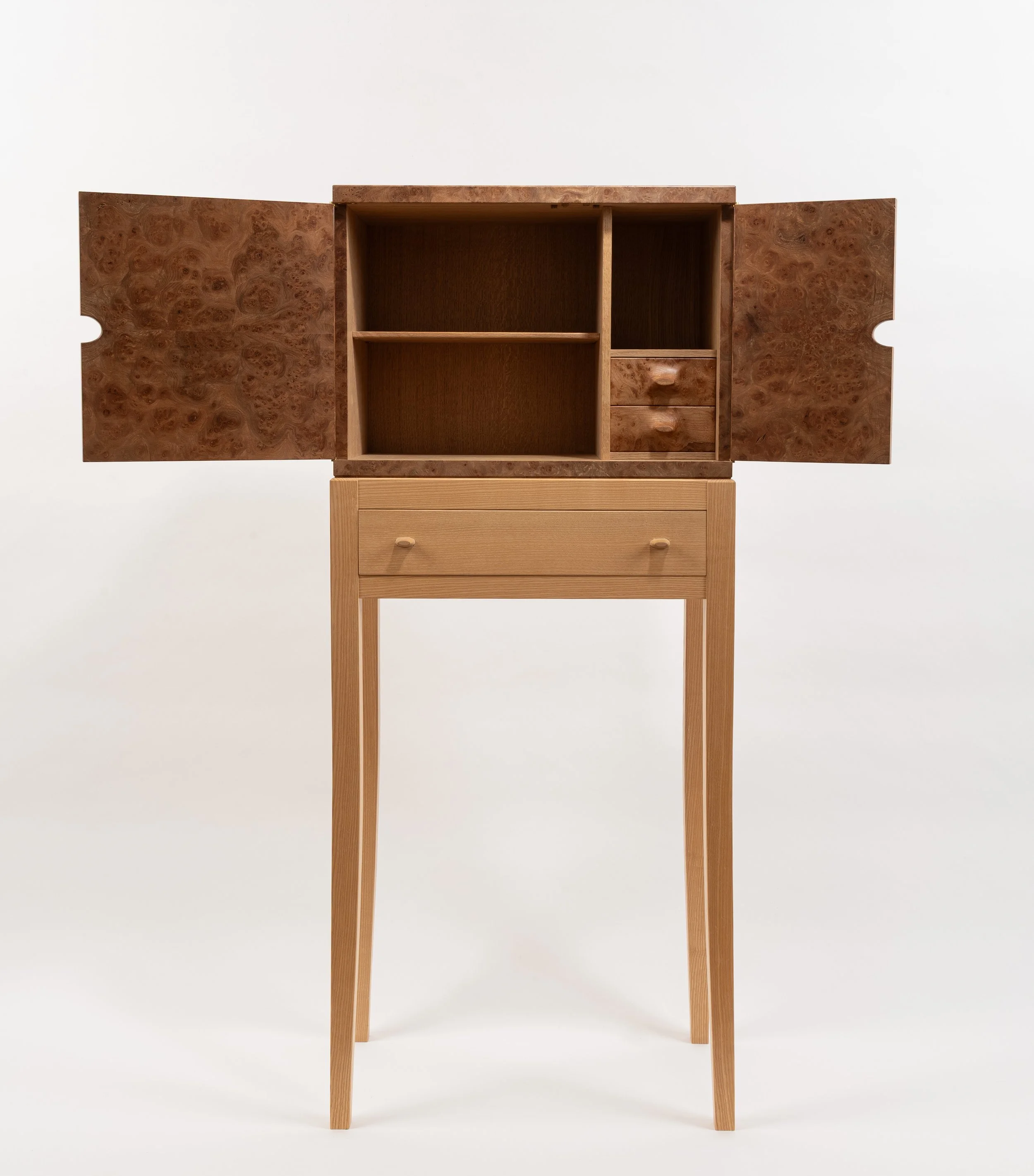 A wooden cabinet with open doors, showing shelves and two small drawers inside, placed on top of a tall wooden table with a drawer.