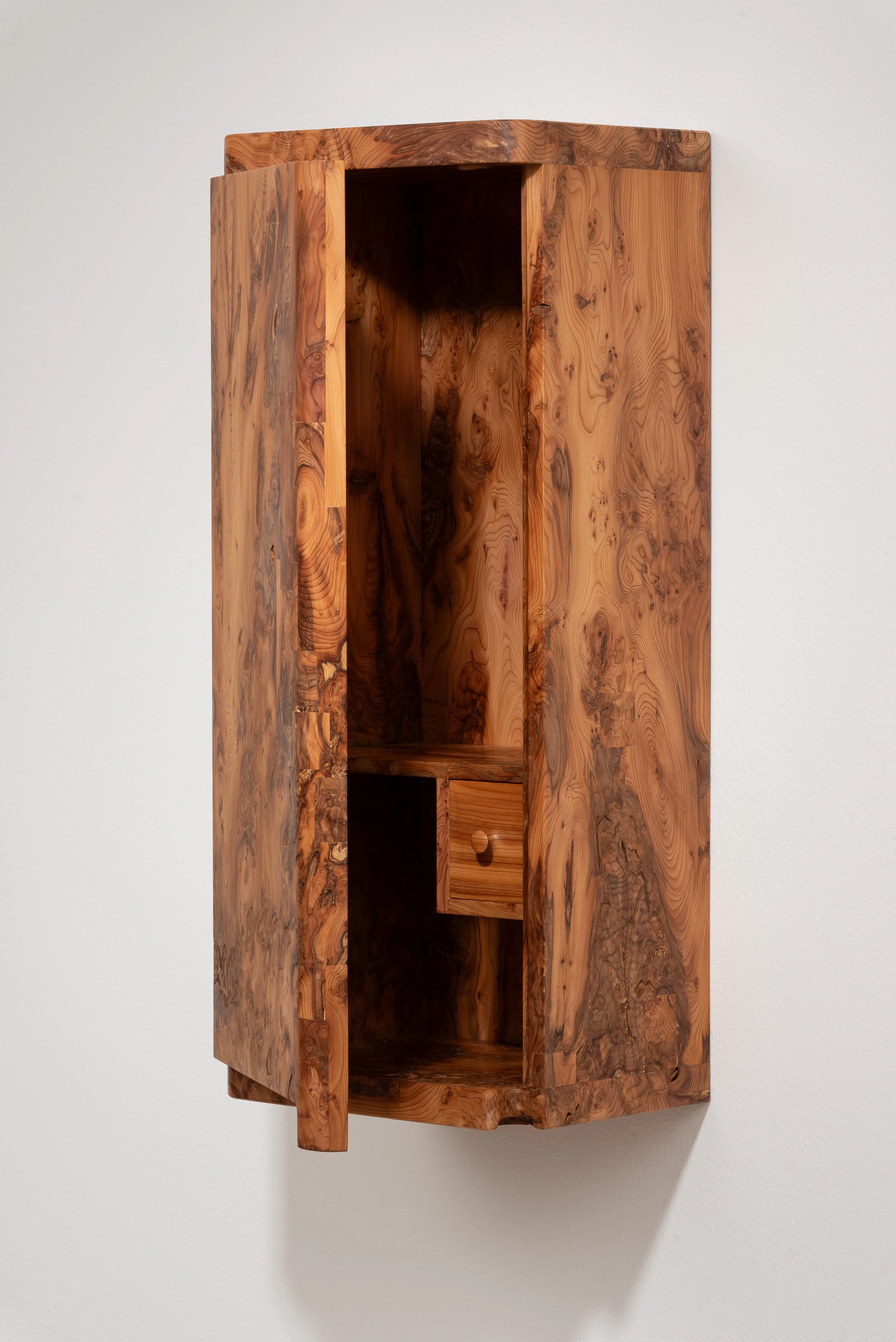 A tall, narrow wooden cabinet with a natural wood finish, mounted on a white wall, featuring a small drawer and open compartments.