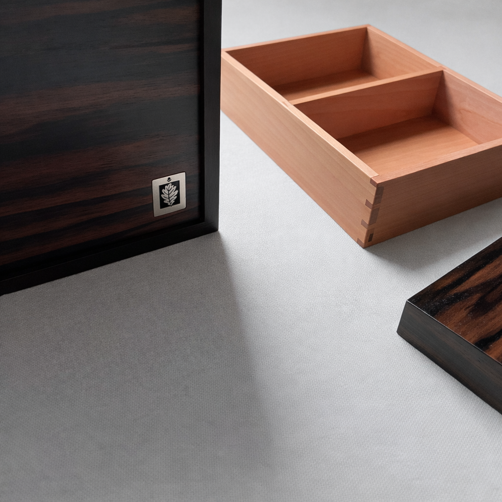 Close-up of a dark wooden cabinet or drawer with a small metallic label, partially open light wooden drawer with a segmented interior, and a dark wooden piece, all placed on a light-colored surface.