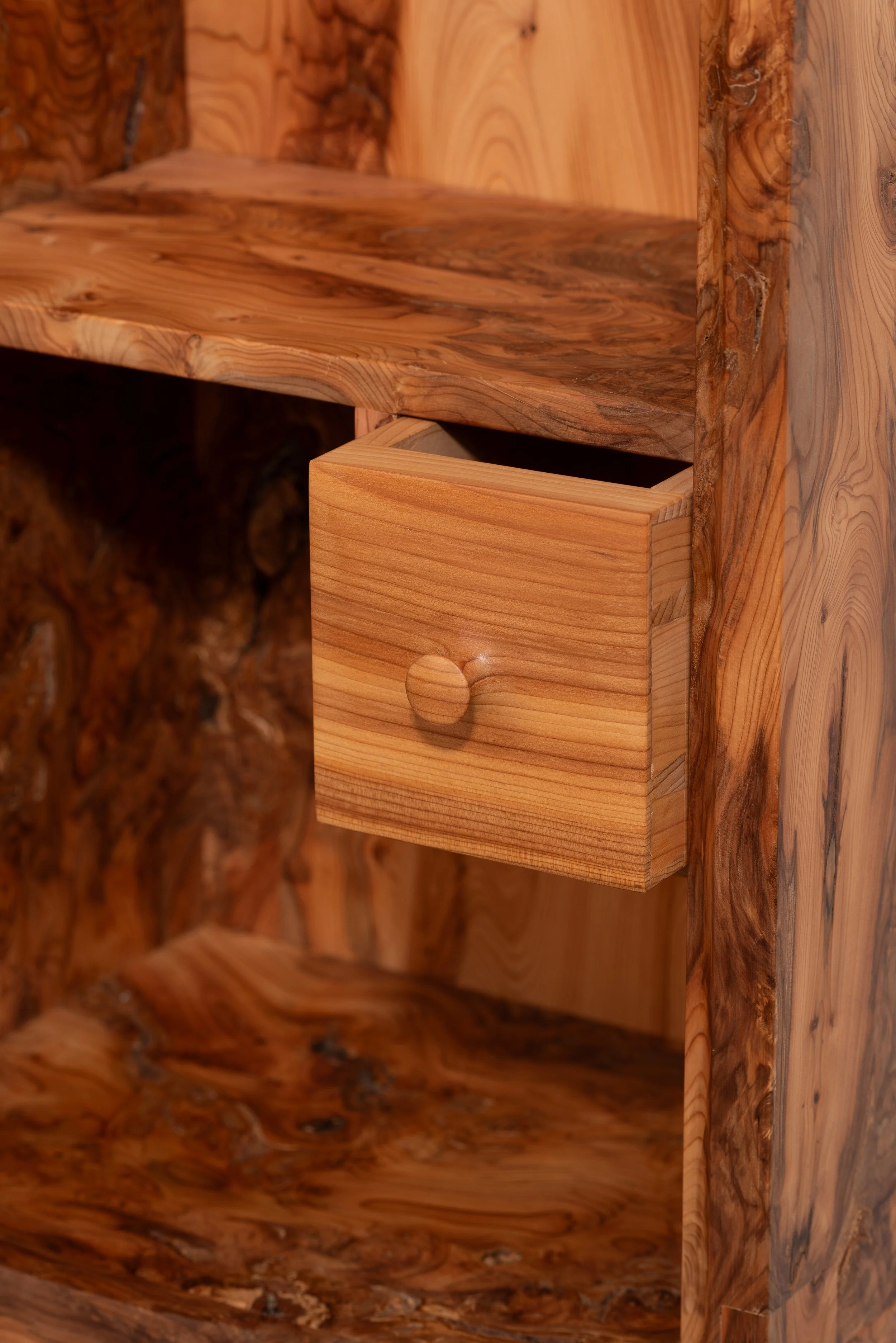 Close-up of a wooden furniture piece with open shelves and a small drawer with a round knob, showing the natural wood grain and knots.