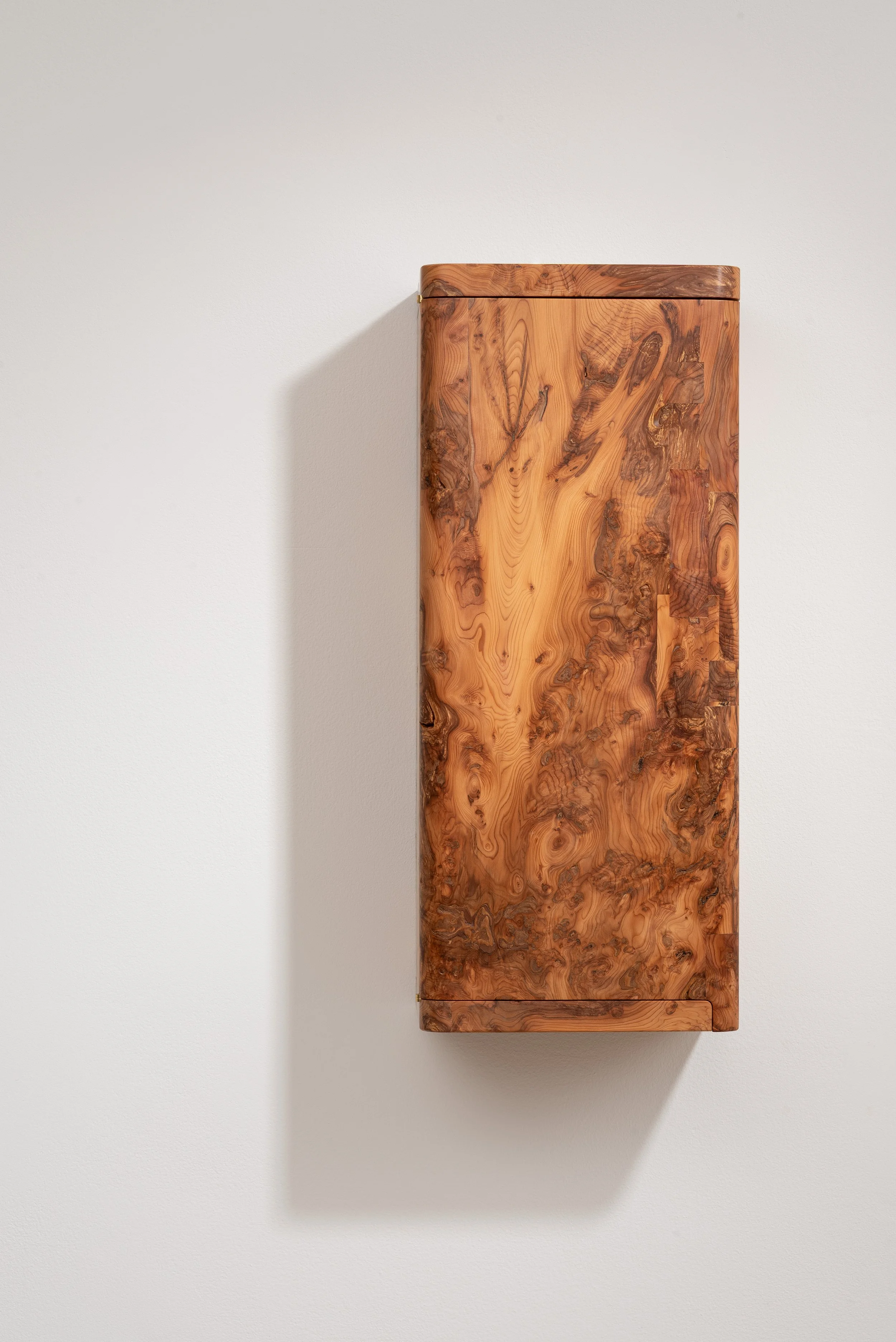 A tall rectangular wooden cabinet with a natural wood grain finish mounted on a white wall, casting a shadow to the left.