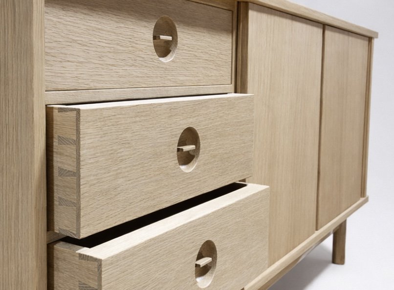 Close-up view of a light wood cabinet with three drawers, each featuring a round cut-out handle, and side panels with horizontal wood grain patterns.