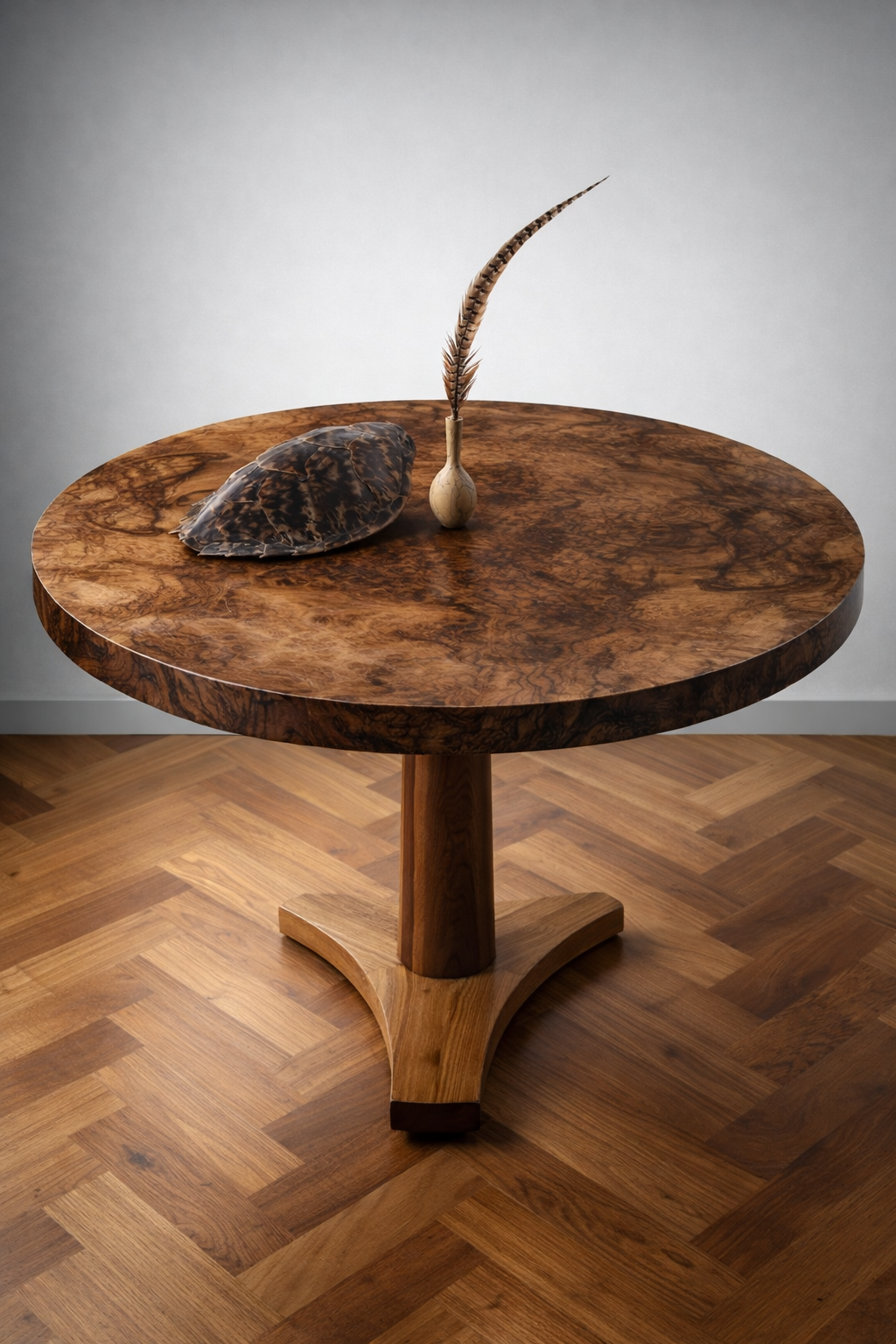 A round wooden table with a polished surface, holding a shell-shaped decorative item, a small vase, and a feather, against a plain gray wall and a wooden floor.