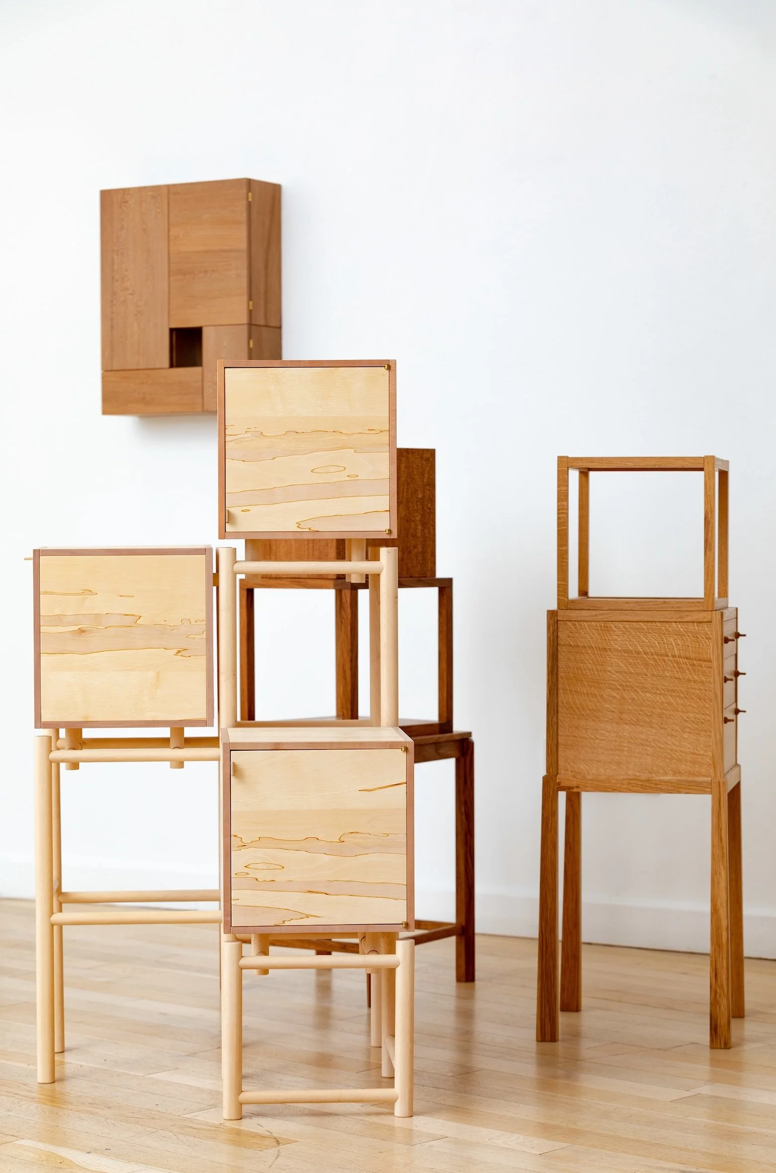 A minimalist display of various wooden furniture pieces, including square and rectangular frames, cubed boxes, and a wall-mounted cabinet, arranged against a white wall on a wooden floor.