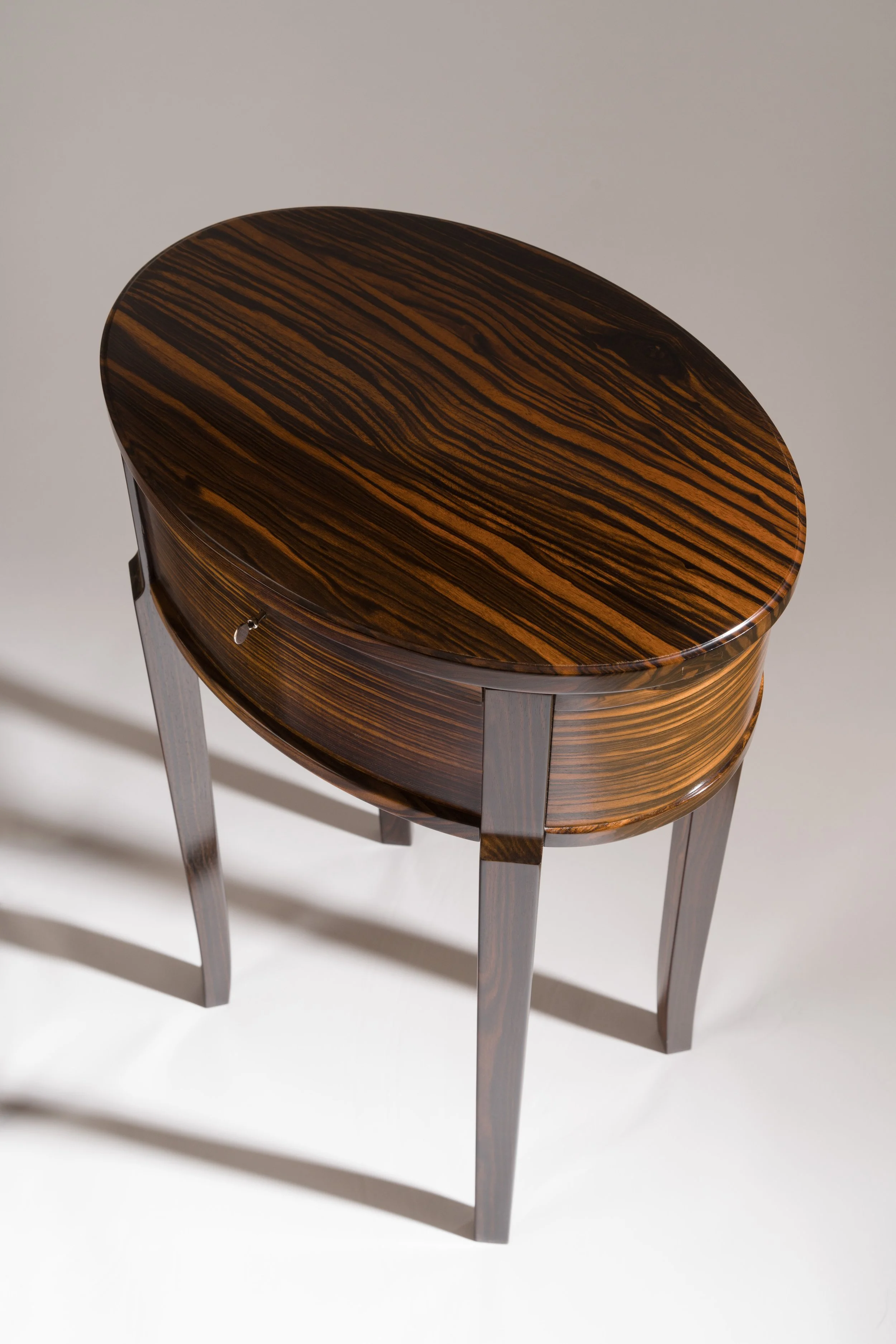 A small, oval-shaped wooden table with a polished, striped wood top and four tapered legs.