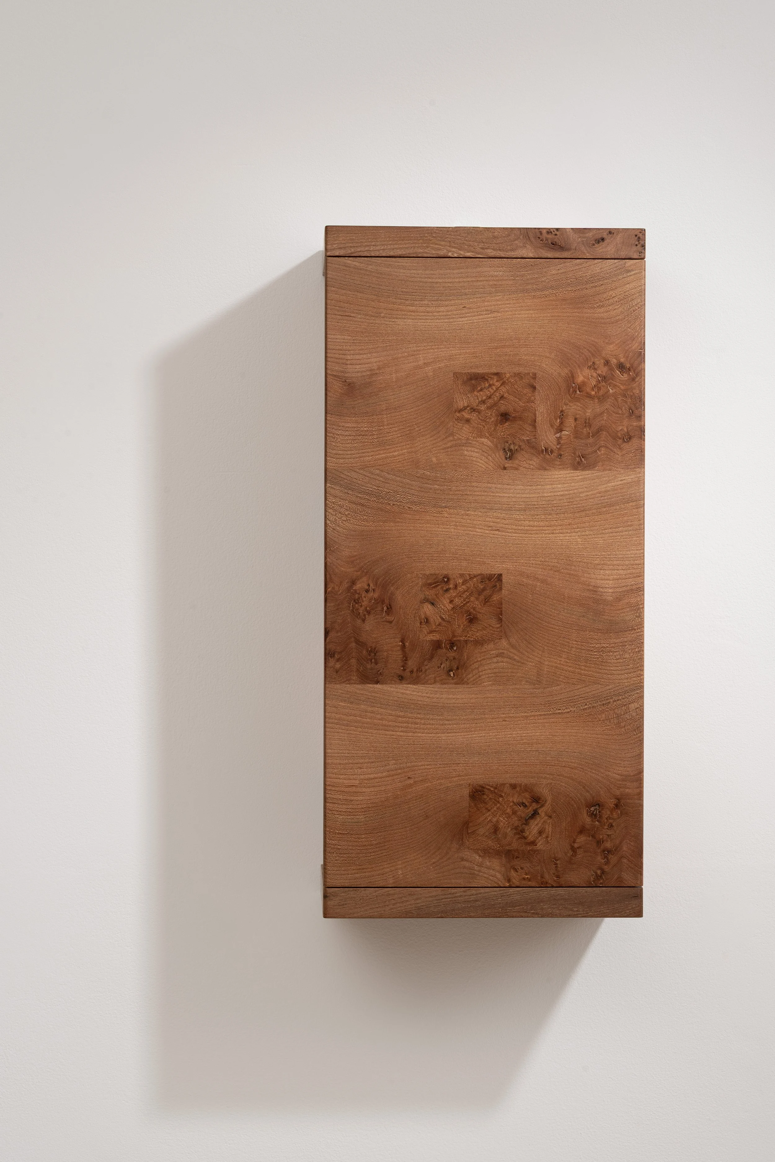 A wooden cabinet with a natural finish mounted on a white wall, casting a shadow.