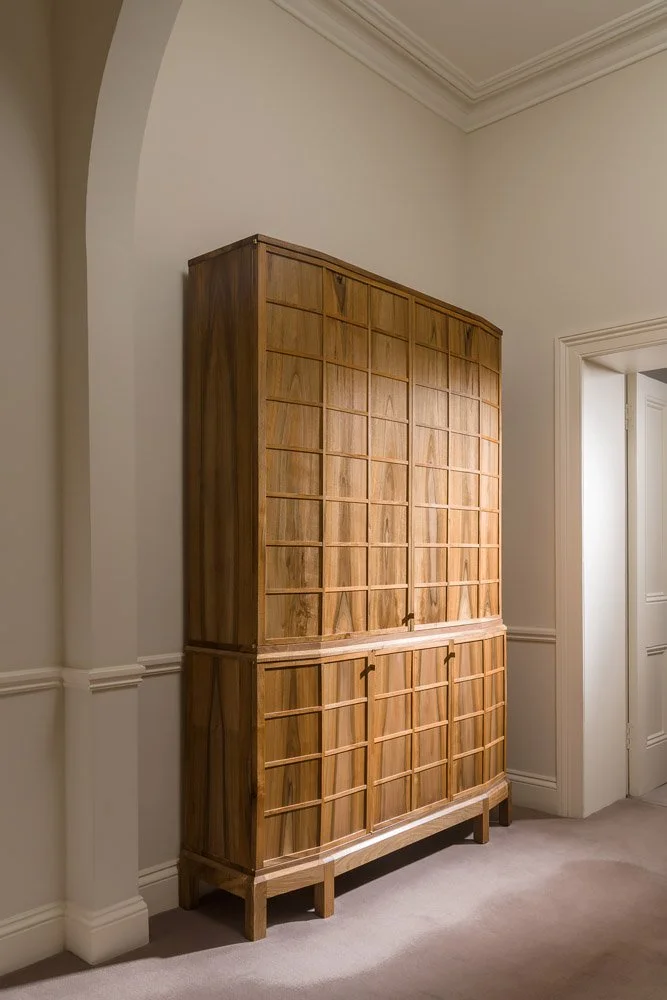 A tall wooden cabinet  in a corner of a room with white walls and beige carpet.