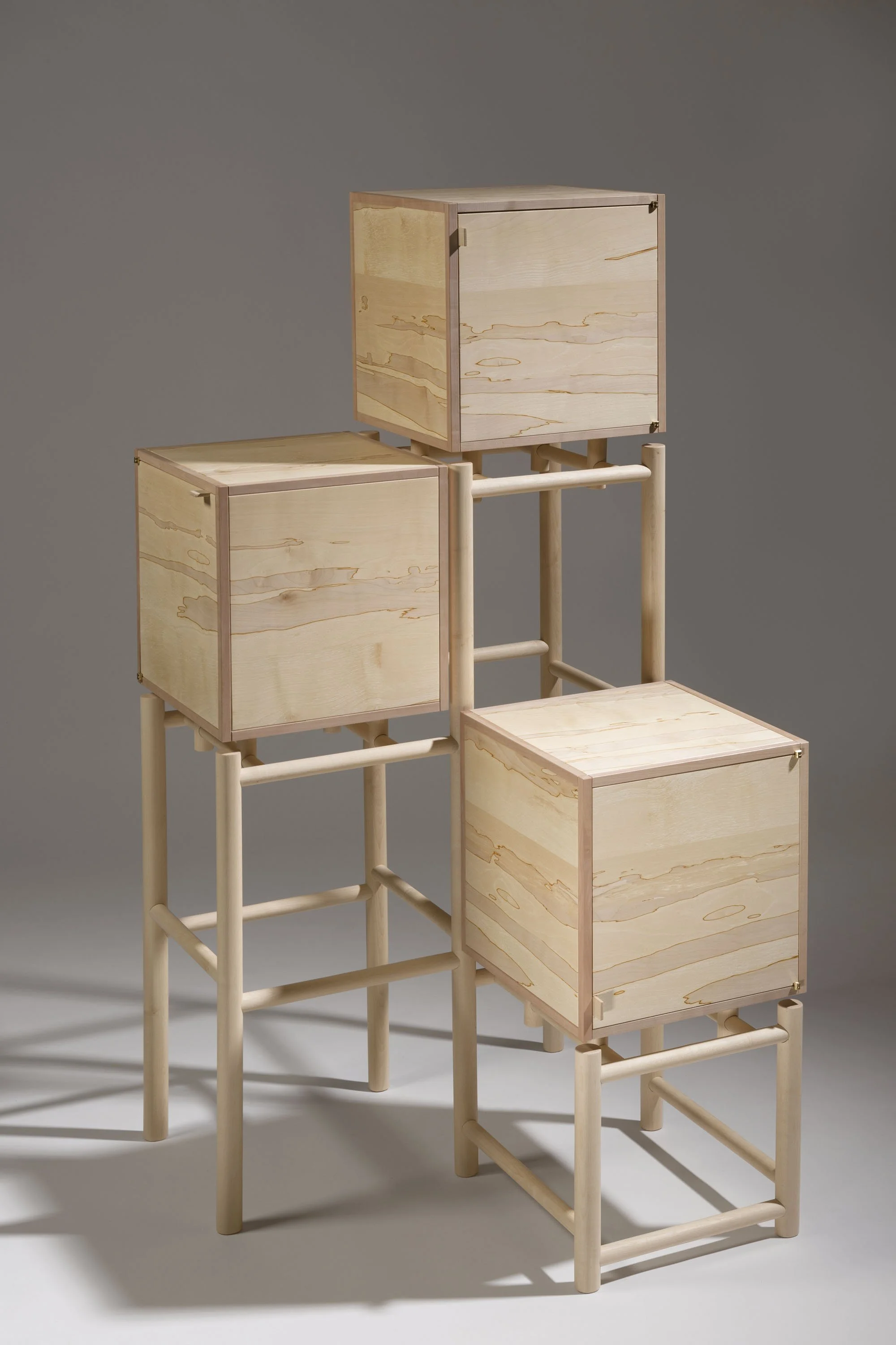 A modern, minimalist cabinet unit with three compartments, made of light-colored wood with a simple frame design, set against a plain gray background.