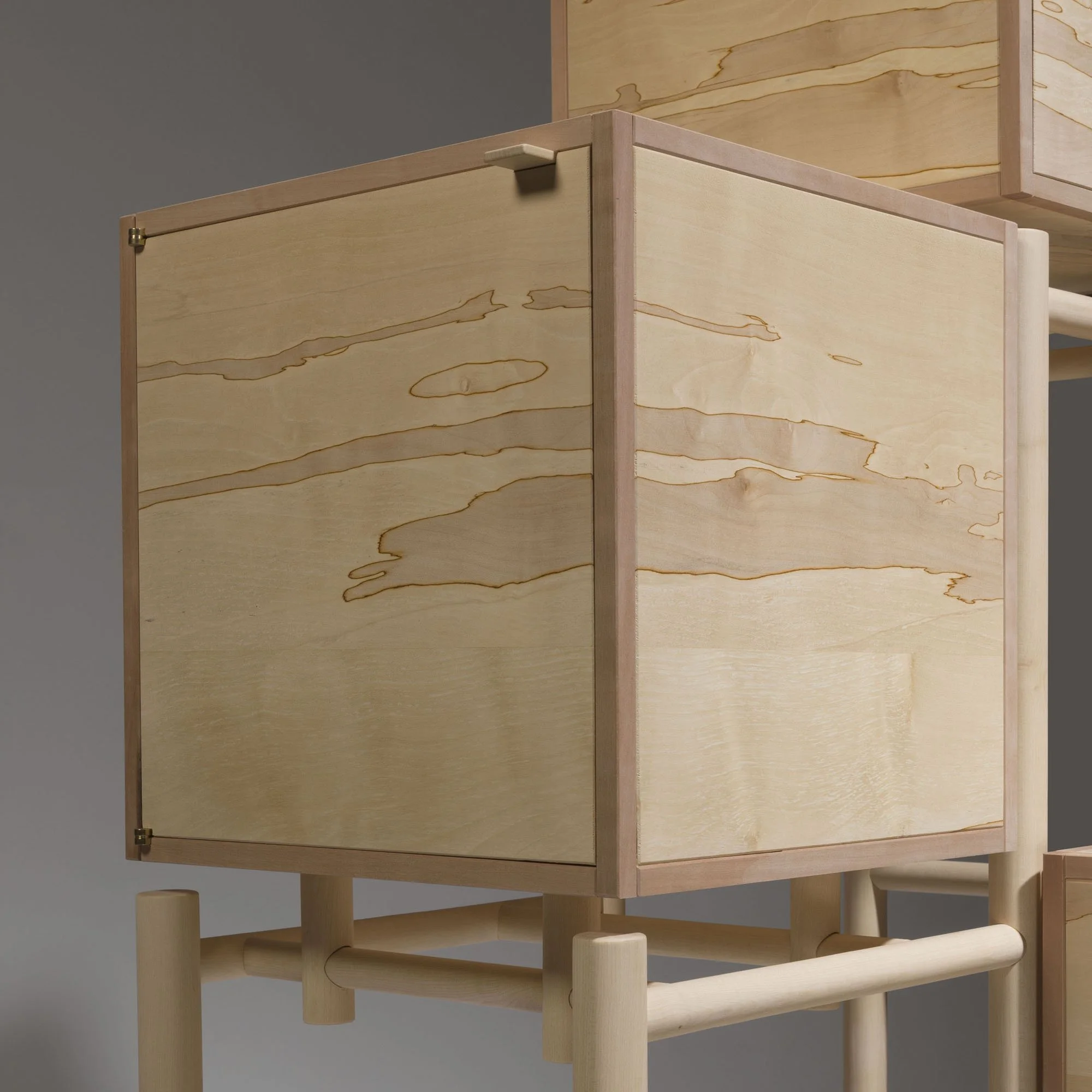 Close-up of a wooden cabinet with a light natural finish and exposed grain, standing on a matching wooden stand with rounded legs, against a plain gray wall.