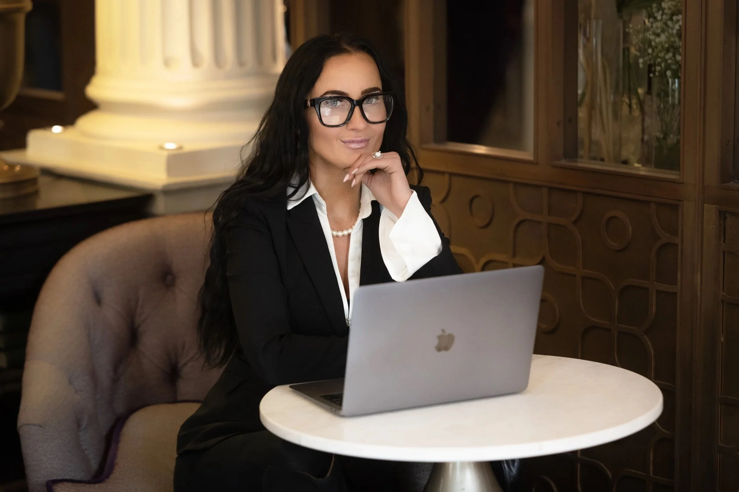 A woman with long dark hair, glasses, and jewelry sitting at a small round table with a MacBook in a cozy, wood-paneled setting.