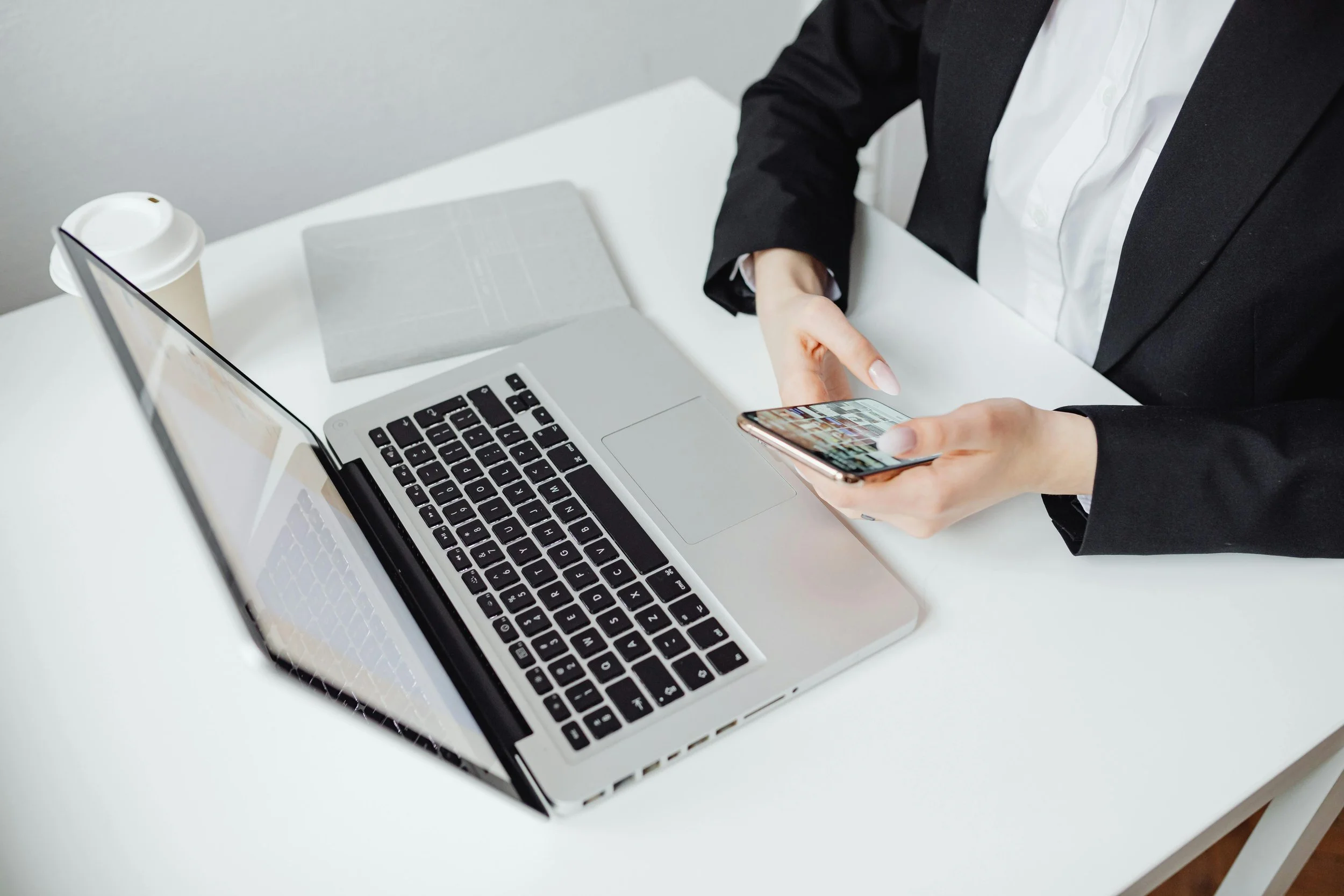 Person in a black blazer using a smartphone at a white desk, with a silver laptop and coffee cup nearby.