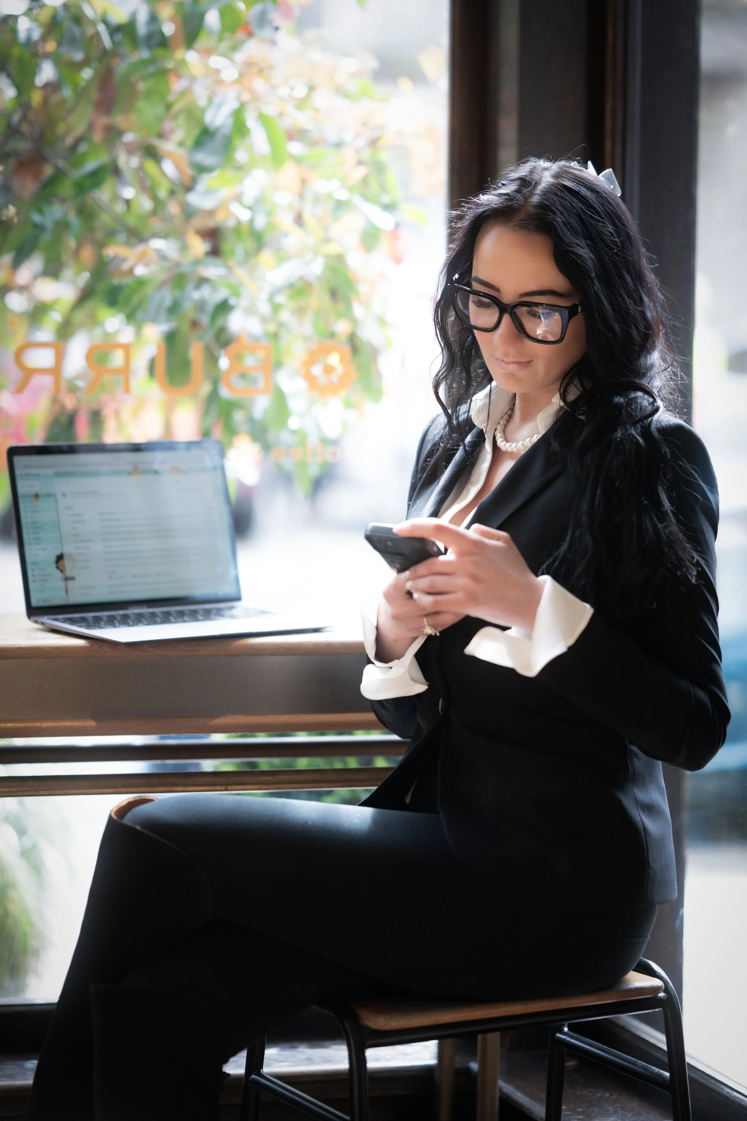 A woman with black wavy hair, wearing glasses, a black blazer, and a pearl necklace, is standing next to a table with a laptop and a smartphone, looking at the phone inside a cafe with large windows and greenery outside.