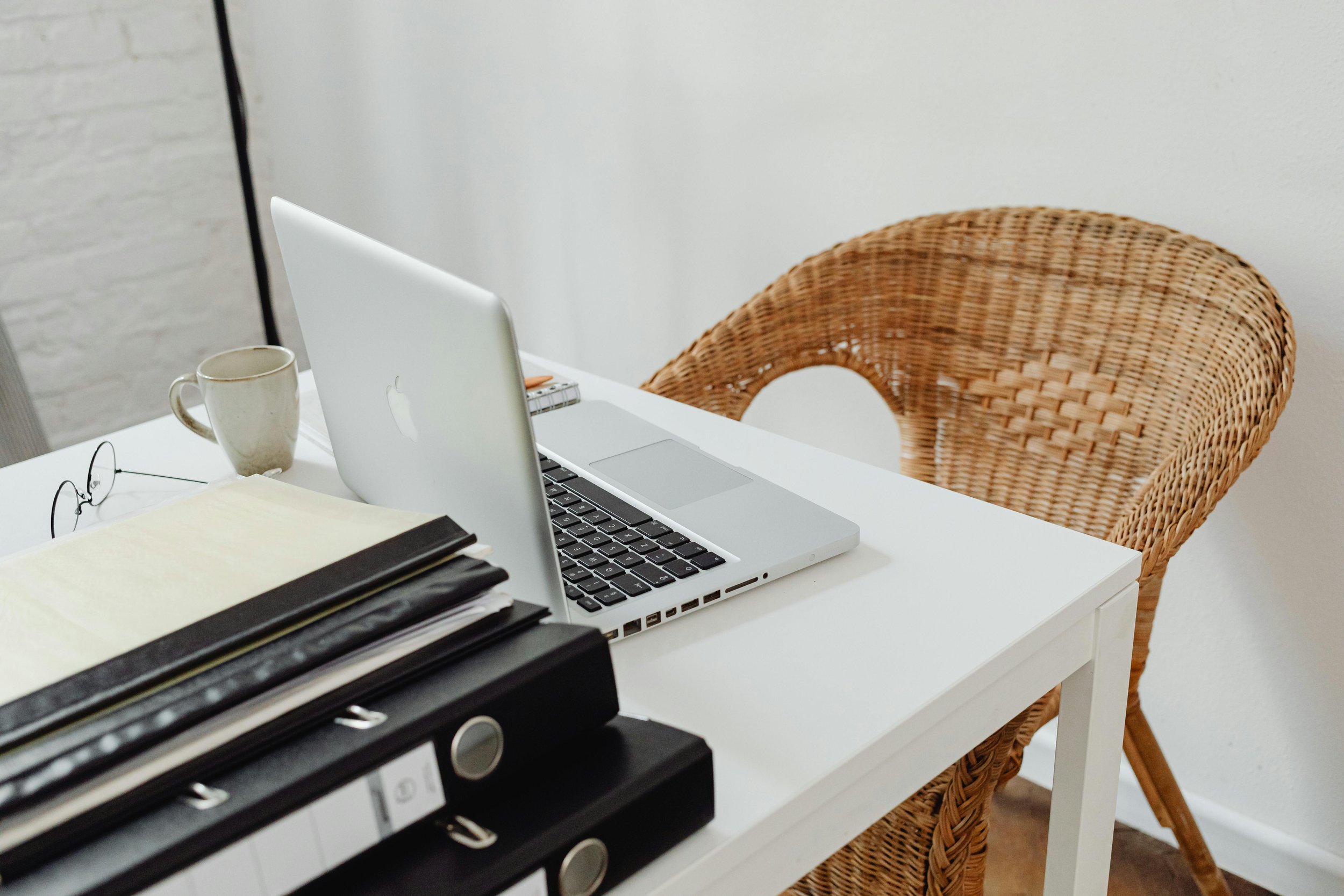 Desk with a closed silver MacBook, a white ceramic mug, glasses, and files in black binders. Wicker chair next to the desk, against a white wall.