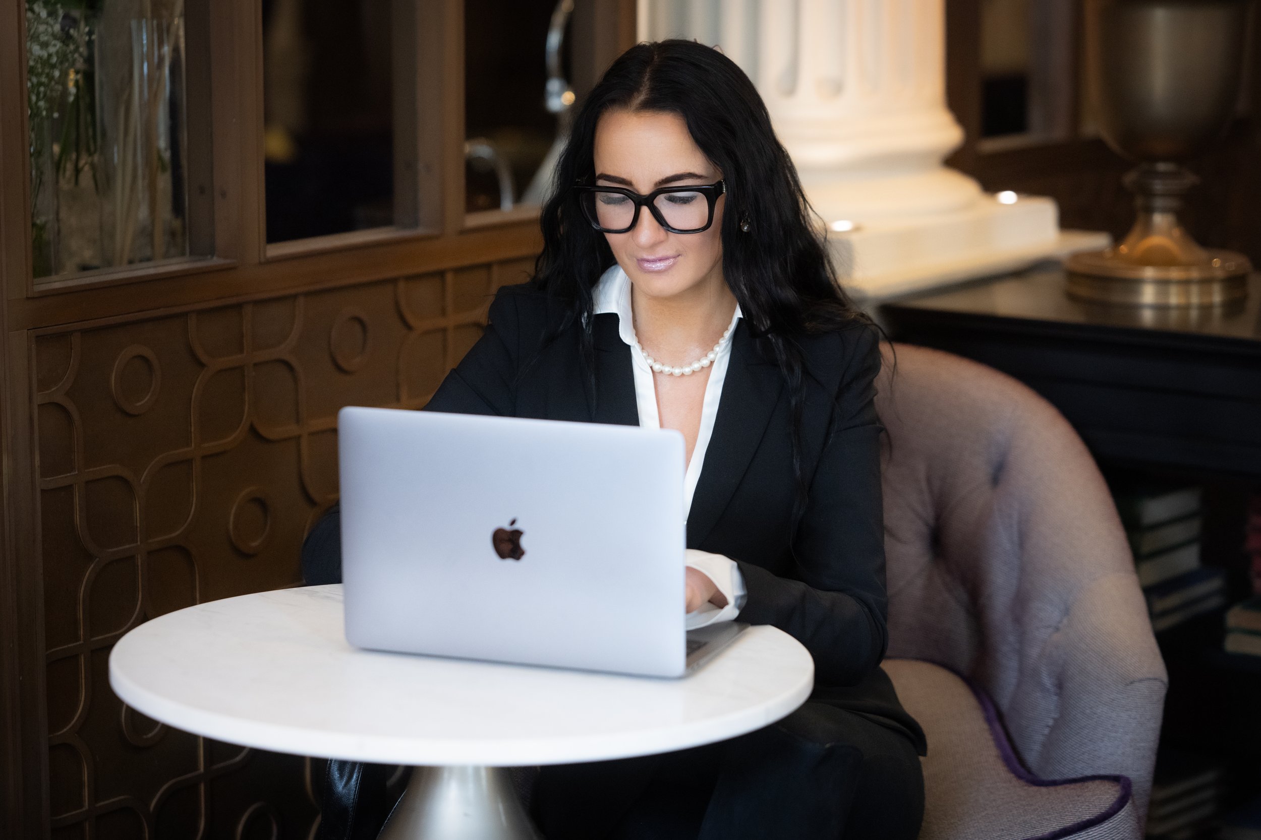 A woman with long black hair, wearing glasses, a pearl necklace, and a business suit, working on a laptop at a small round table in a cozy, elegant cafe or restaurant.