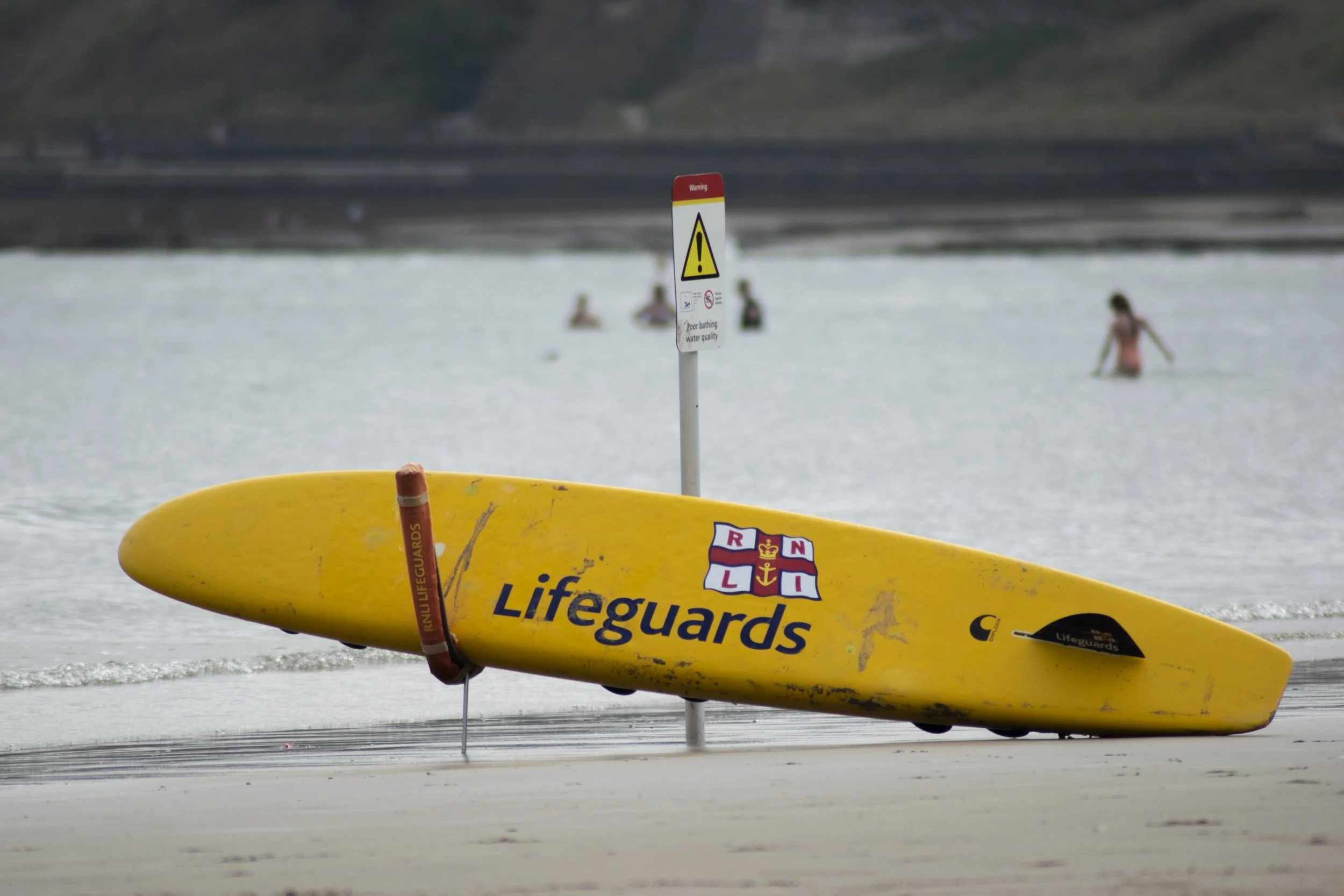 Yellow lifeguard surfboard resting on the sand near the water with a warning sign nearby, and several people in the water in the background.