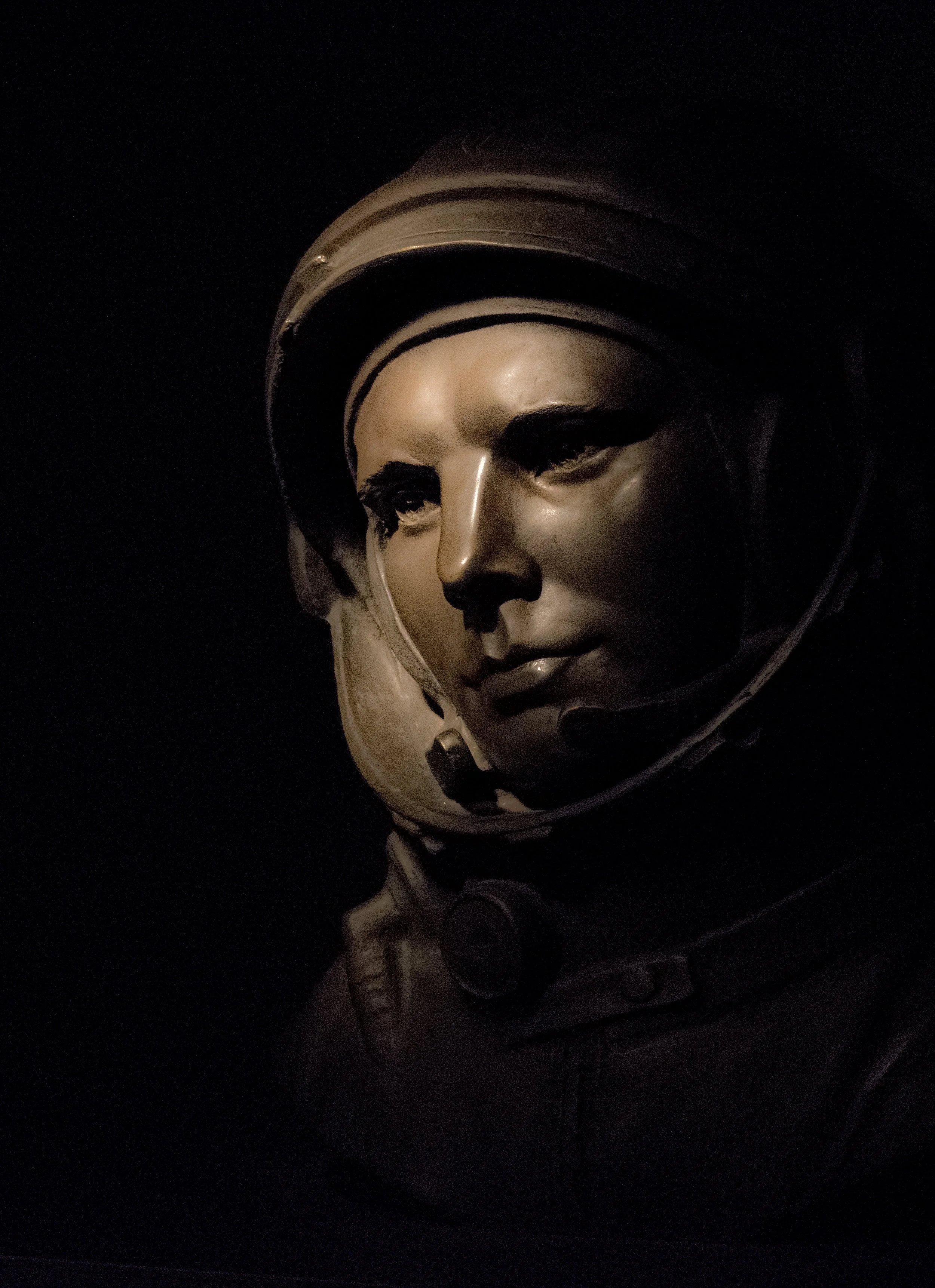 A close-up of a mannequin head wearing a vintage astronaut helmet, with shadowed face and minimal lighting highlighting the helmet's structure.