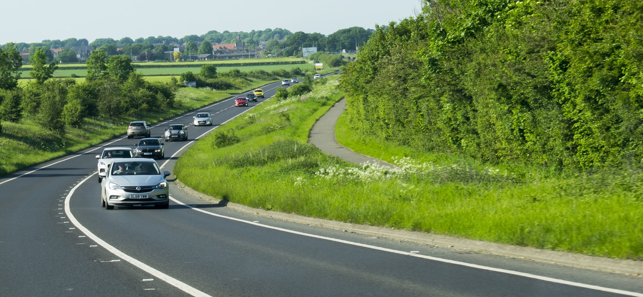 Cars driving on a curving two-lane highway through lush green countryside with trees and grass along the sides and a small sidewalk on the right.