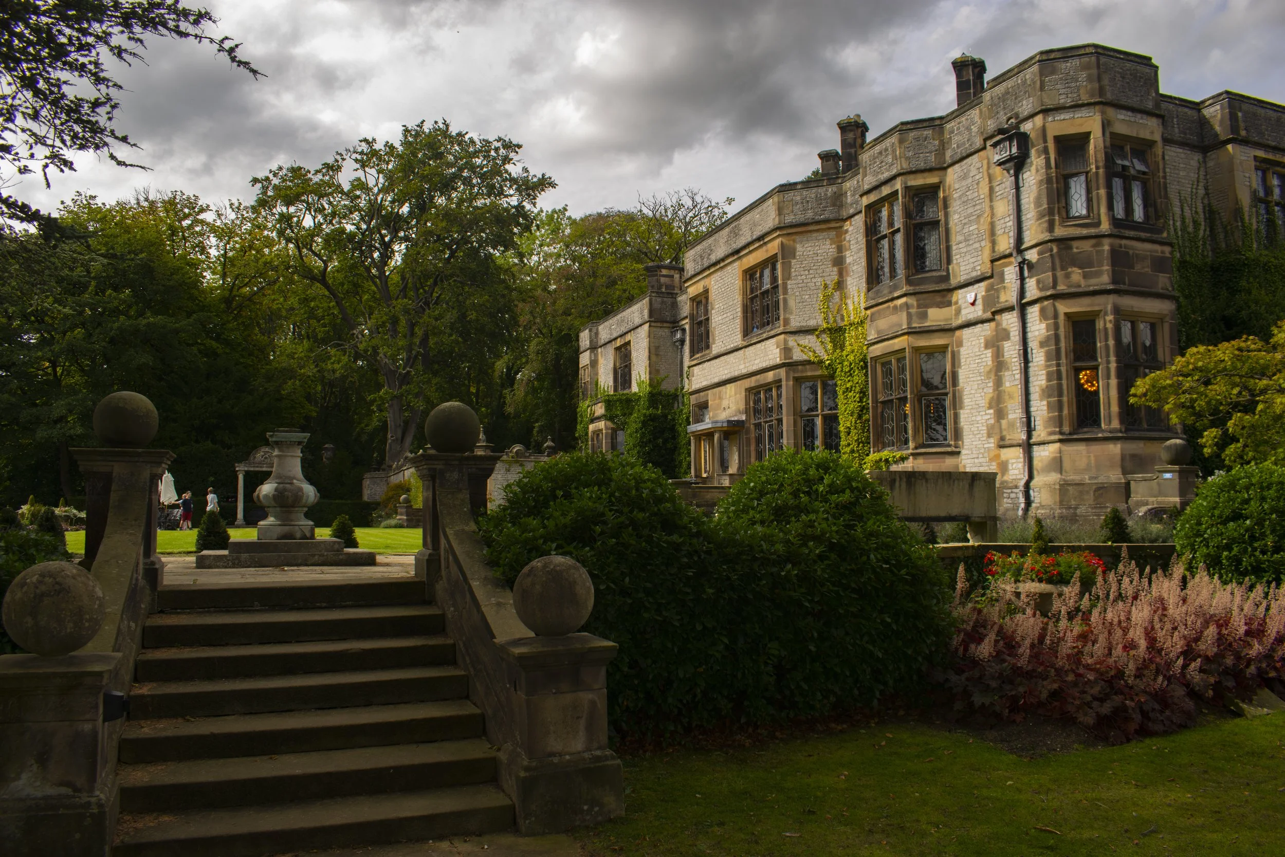 A large stone mansion surrounded by lush greenery and well-maintained gardens, with a stone staircase leading up to the house on an overcast day.