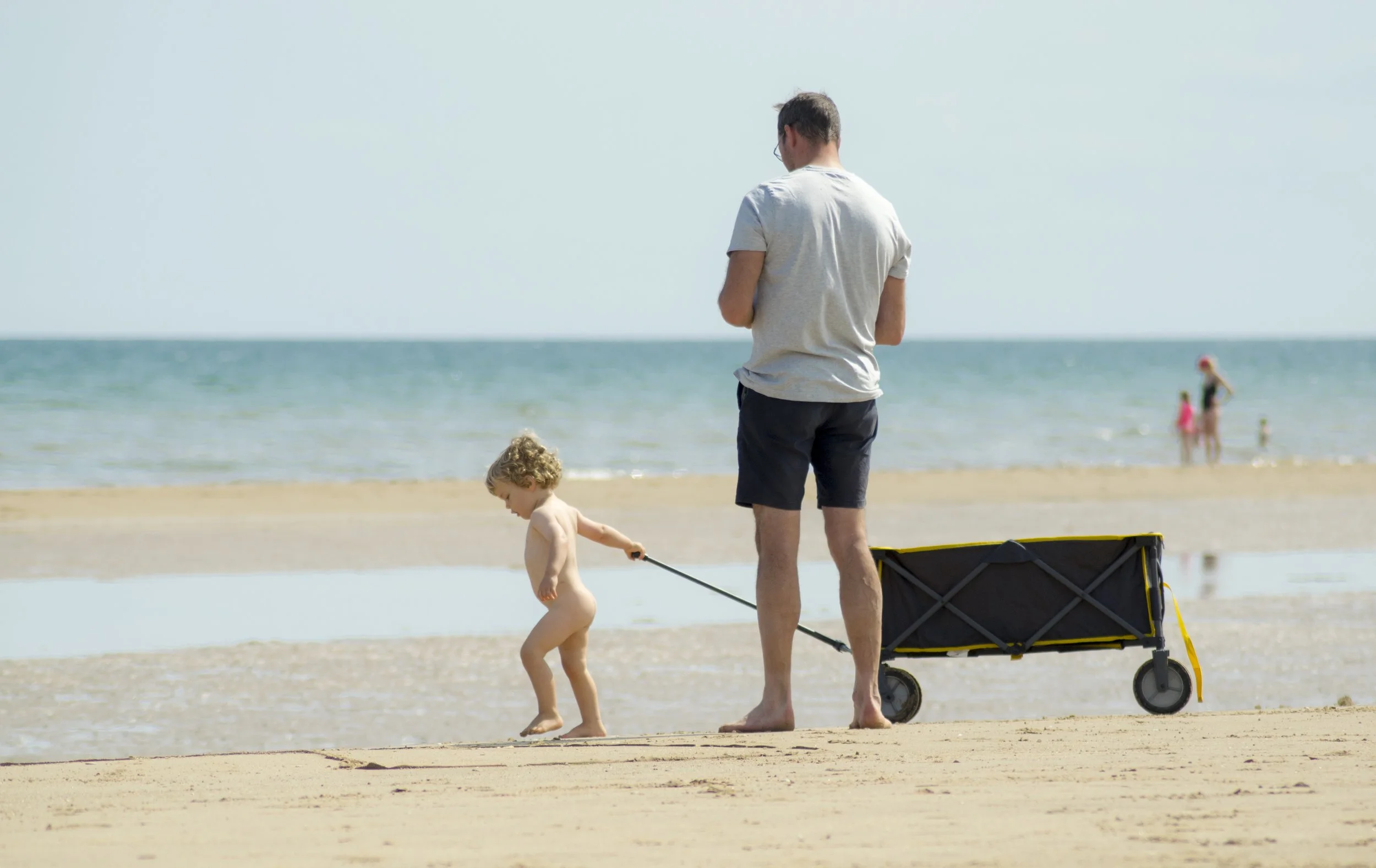 A man walking on the beach pulling a small wagon while a young child, who is not wearing clothes, holds the wagon's handle. In the background, the ocean and a woman with children are visible.