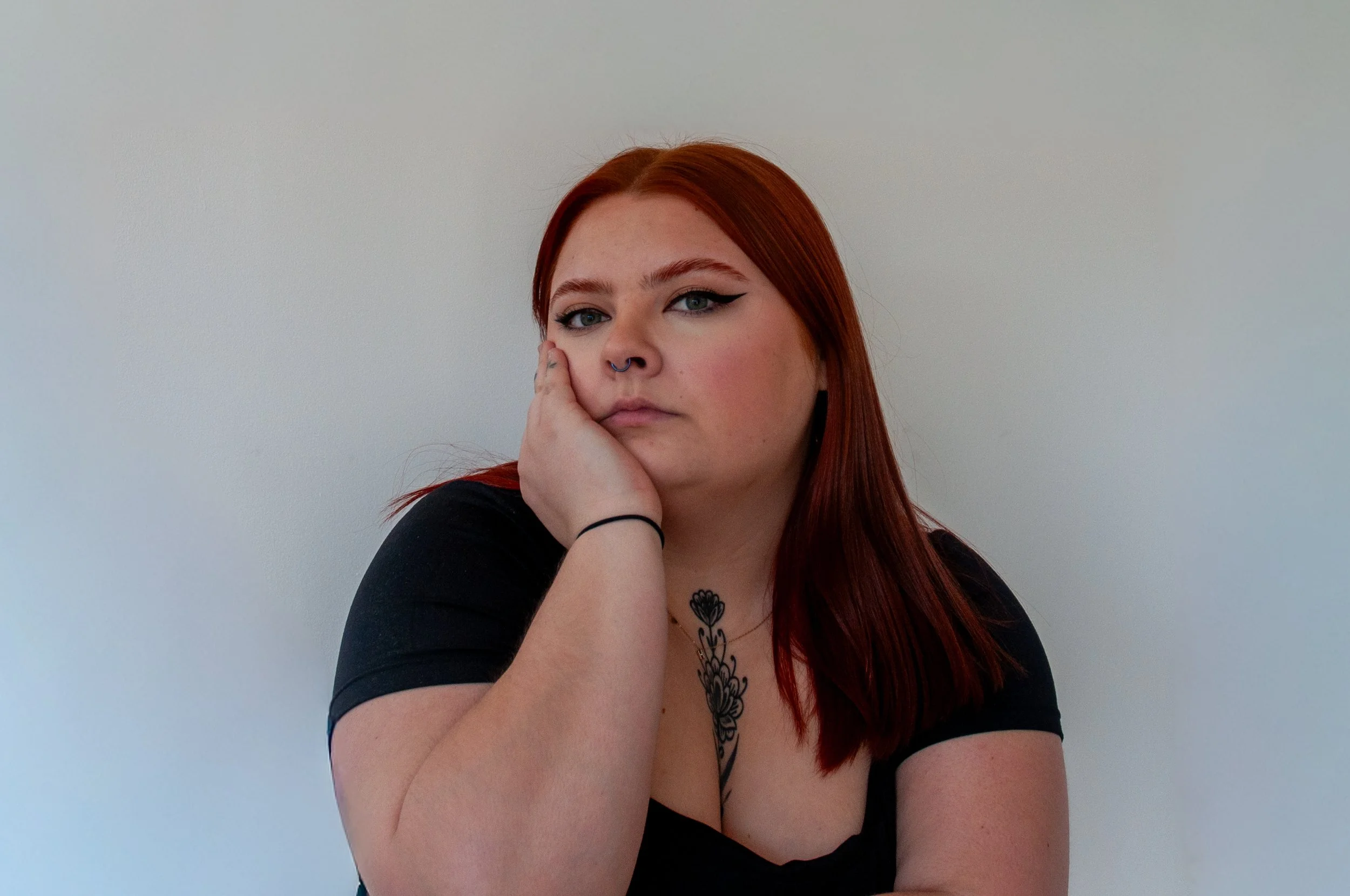 A young woman with red hair and a septum piercing resting her head on her hand, wearing a black top, with a visible floral tattoo on her chest, against a plain white wall.