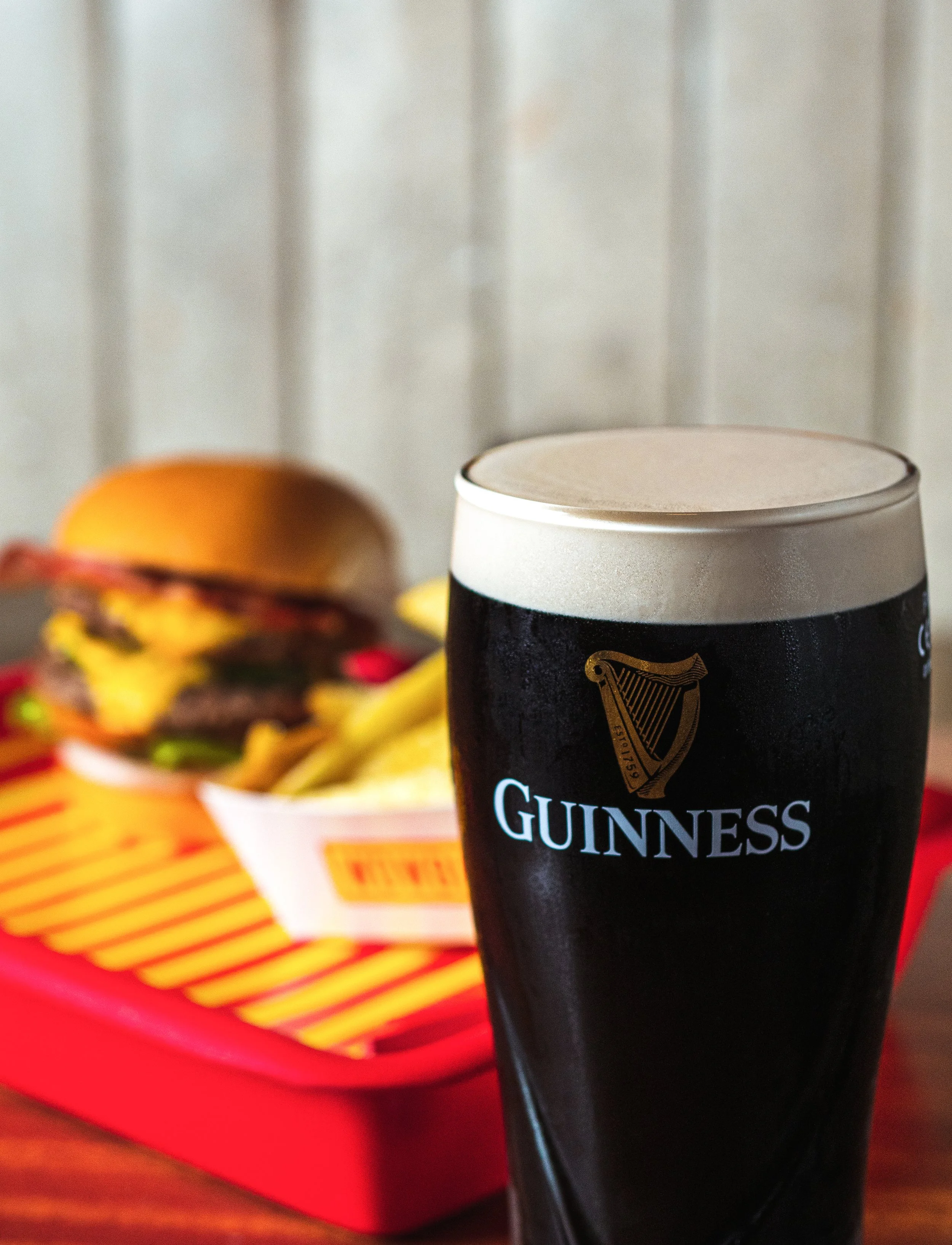 A pint of Guinness beer in the foreground with a tray of fast food including a burger and fries in the background.