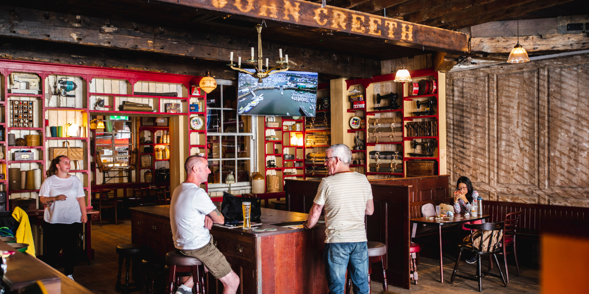 People inside a rustic bar or pub with wooden beams, red and white shelving, and a television mounted on the wall, some sitting and one standing near the bar.
