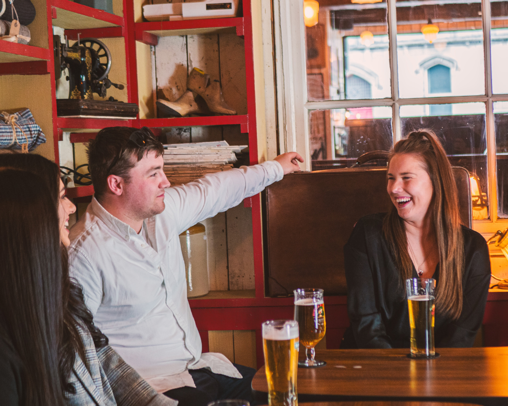 Three friends sitting at a wooden table in a cozy restaurant or pub, enjoying drinks and conversation, with a red shelving unit behind them and a window showing the street outside.