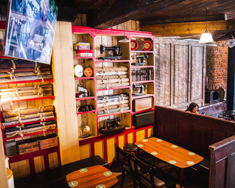 Interior of a cozy restaurant with wooden walls and ceiling, decorative shelves displaying vintage sewing machines and fabric rolls, and tables set with coasters. A person is seated at a table near a brick wall, and a large television is mounted on a