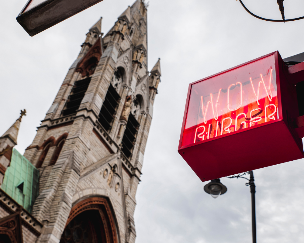 A neon sign that reads 'WOM' with the word 'BURGER' underneath, hanging outside near a tall church in the background.