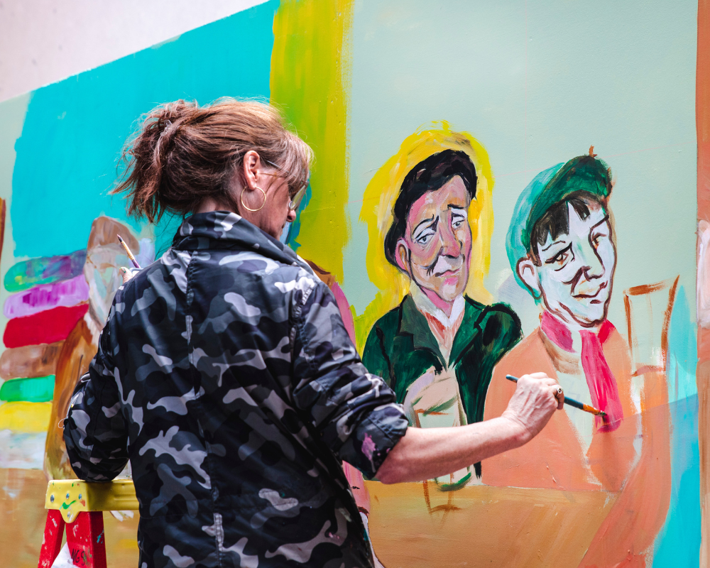 An artist painting a colorful murals of two women with expressive faces.