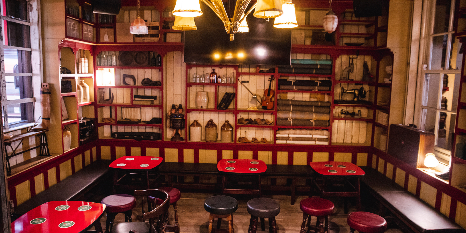 Cozy restaurant interior with red and black tables, wooden chairs and benches, and a wall with shelves filled with books, bottles, instruments, and decorative items, illuminated by warm lighting and lamps.