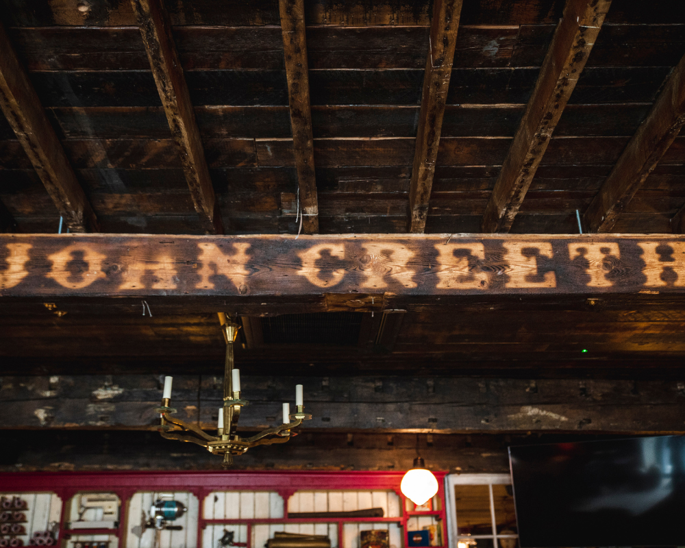 Wooden beam with the words "NO ENTRY" stenciled on it in black, hanging under a rustic wooden ceiling in a bar or restaurant setting.