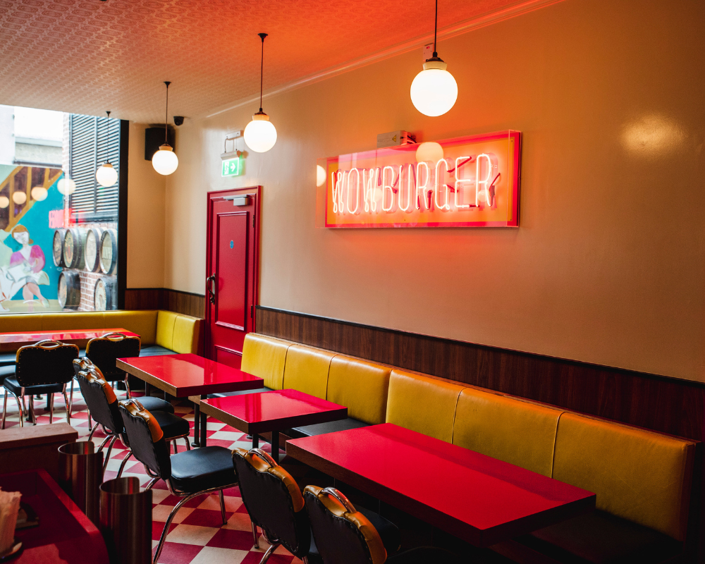 Interior of a burger restaurant with yellow booths, red tables, black chairs, and a neon sign that says 'Hamburger' on the wall.