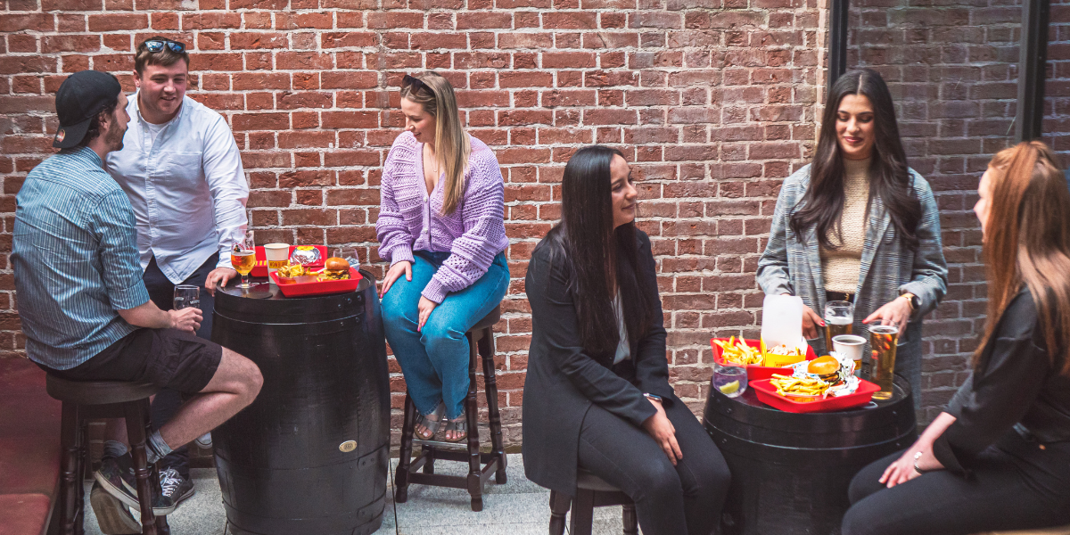 Six young women and men socializing outside near a brick wall. They are sitting around two black barrels that hold snacks and drinks, including burgers, fries, and beers.
