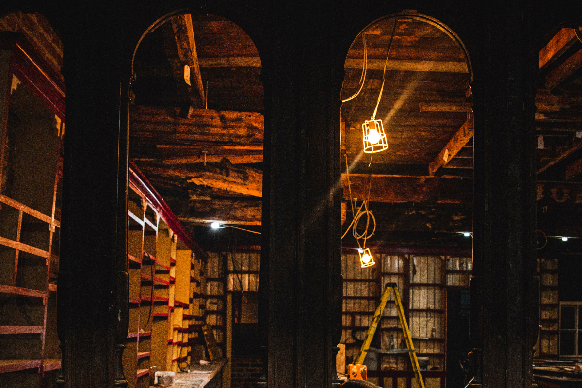 Interior of a building under construction or renovation, with exposed wooden beams on the ceiling, a ladder, and hanging industrial-style lights.