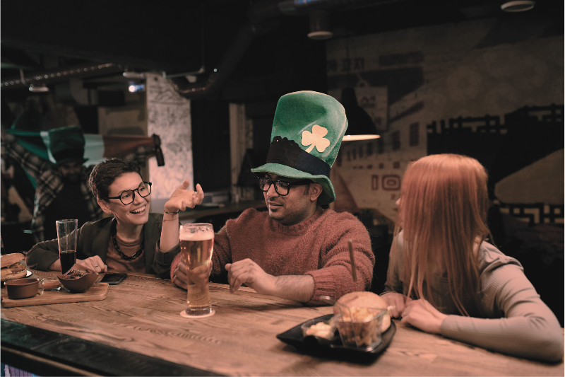 Three friends sitting at a bar, one wearing a large green St. Patrick's Day hat with a shamrock, enjoying drinks and conversation.