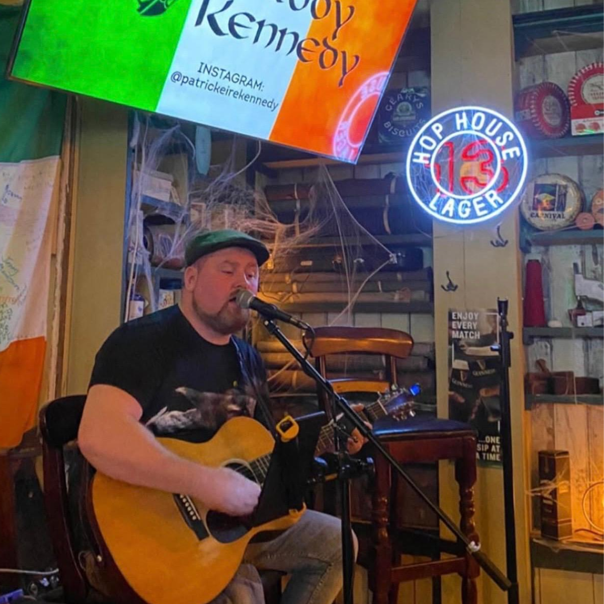 A man wearing a green cap and black T-shirt, playing an acoustic guitar and singing into a microphone in a pub decorated with Halloween cobwebs.