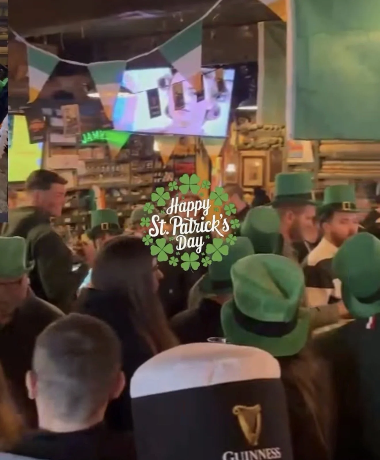 Crowd at a bar celebrating St. Patrick's Day, many wearing green hats, with a sign that says 'Happy St. Patrick's Day' and shamrock decorations.