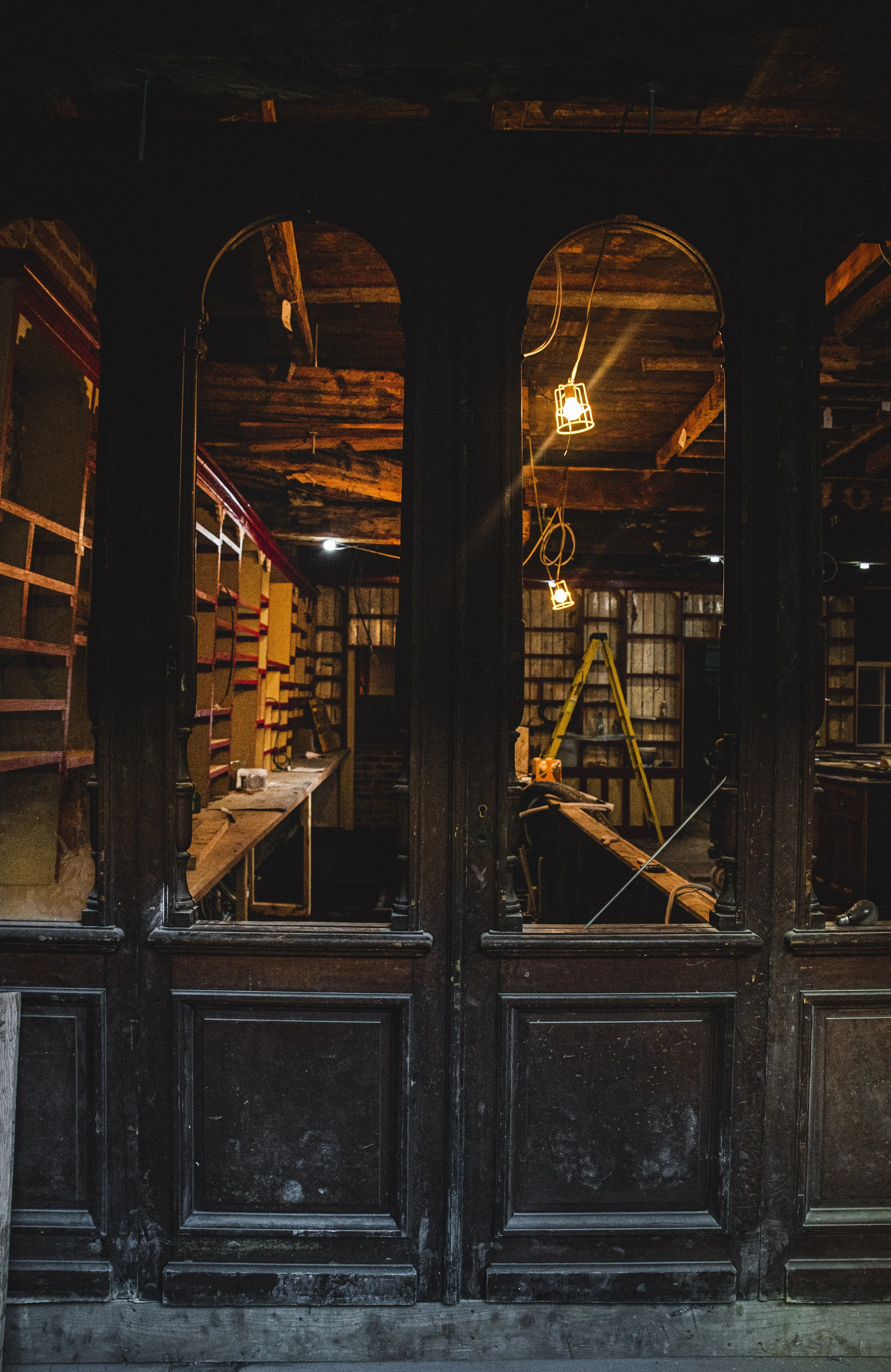 Interior of a rustic, unfinished woodworking shop with dark wooden framing, shelves, ladder, and hanging industrial-style lights.