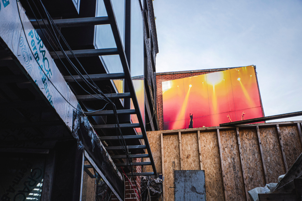 Construction site with a partially built brick wall, wooden framing, and a large billboard showing a stage with bright yellow and orange lighting.