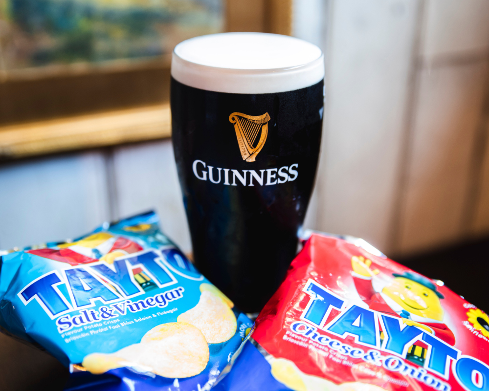 A glass of Guinness stout beer with a foamy head, placed in front of two bags of TAYTO potato chips, one salt & vinegar flavor and the other cheese & onion flavor.