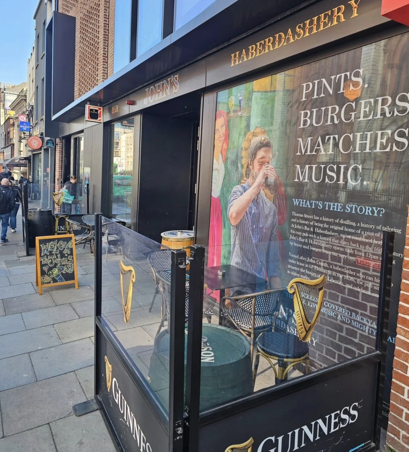 Street view of a pub with a large window display showing people enjoying drinks, a poster with Irish symbols and text about the pub's history, outdoor seating, and a chalkboard sign with welcoming message.