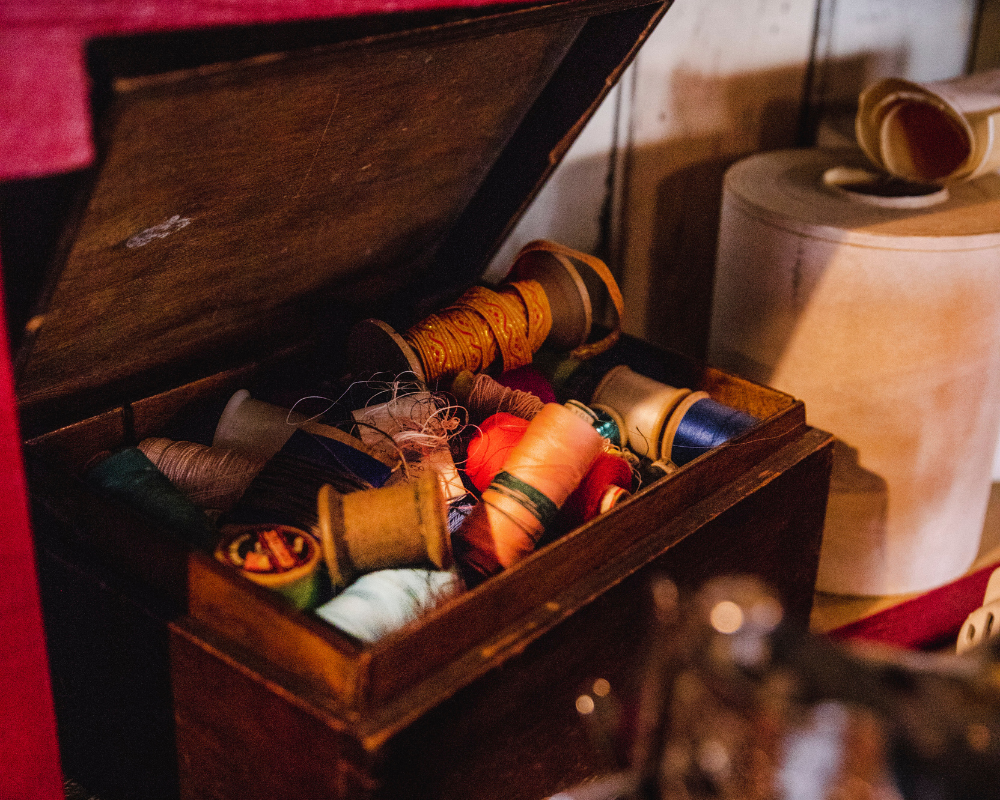 An open wooden box filled with colorful spools of thread and sewing supplies. In the background, a cardboard box and a roll of paper or fabric are visible.