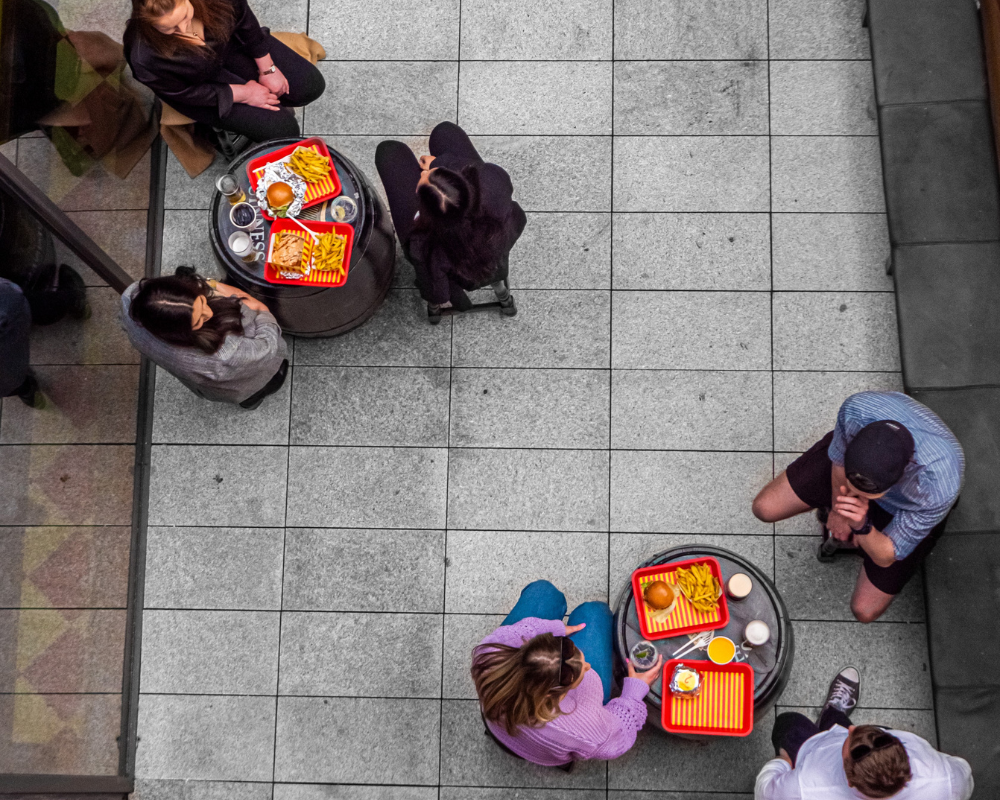 Top-down view of two tables with meals and people, surrounded by five people sitting or standing on tiled flooring.