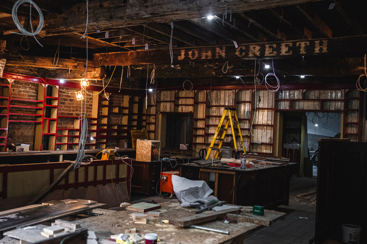 Interior of a restaurant or bar under renovation with exposed brick walls, an unfinished ceiling with hanging wires, a yellow ladder, and construction tools on a cluttered work table.