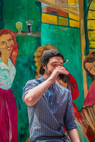 Man wearing a baseball cap and striped shirt drinking soda in front of a colorful mural.